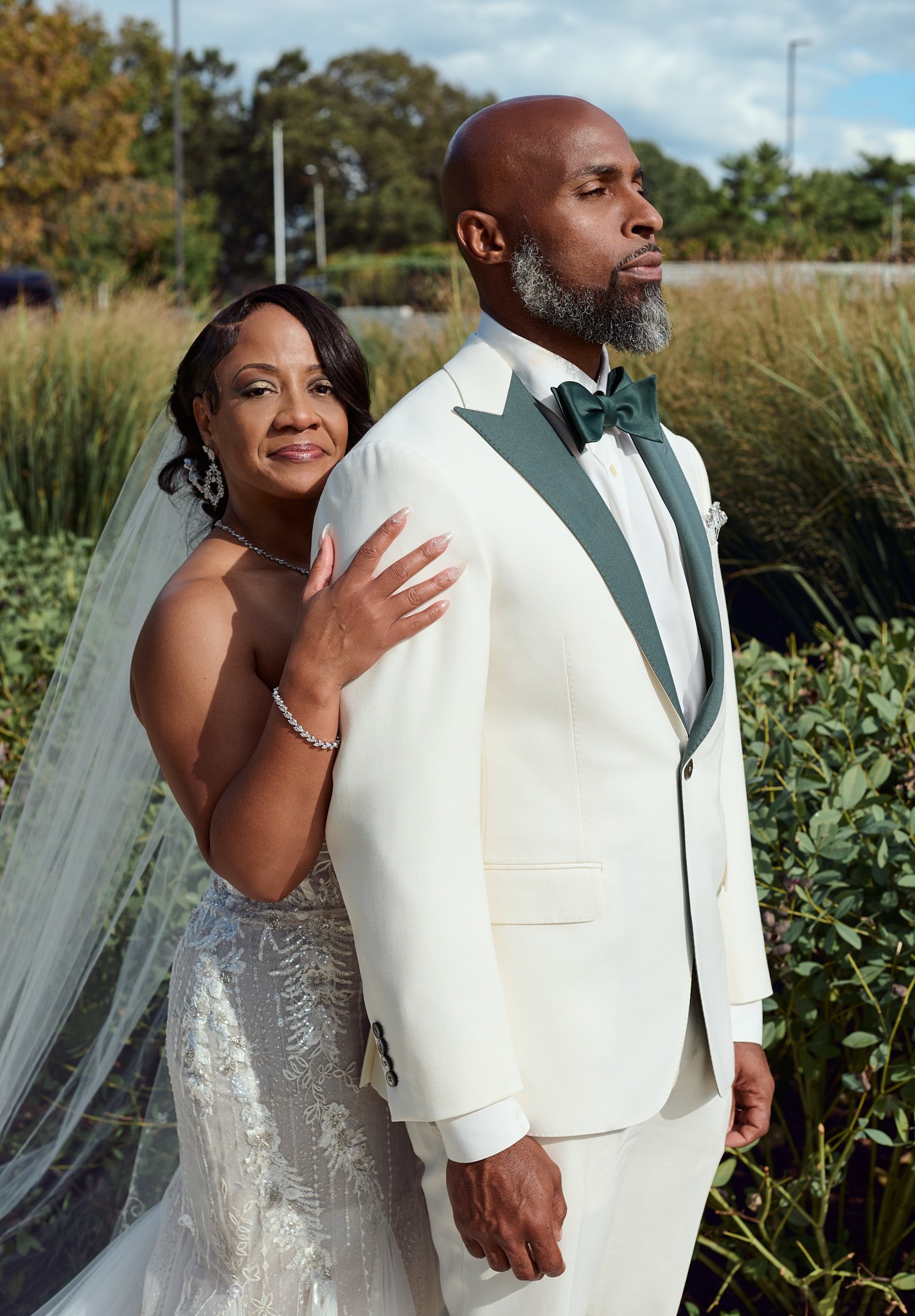 A bride and groom posing outdoors, with the bride resting her hand on the groom's shoulder. The groom is dressed in a white tuxedo with a dark green bow tie and dark green lapels, and the bride is in a strapless lace wedding gown with jewelry. There 