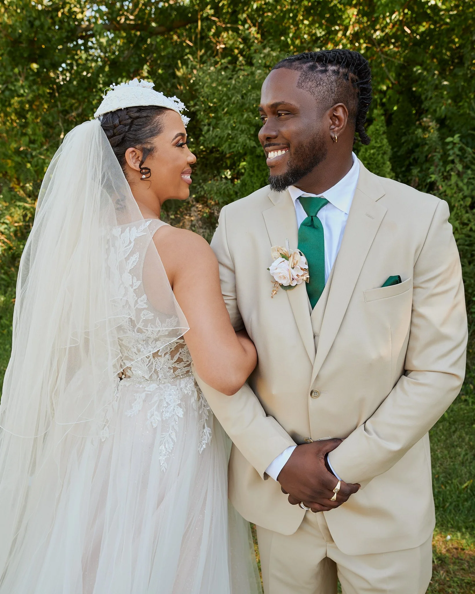 A bride and groom standing outdoors, facing each other and smiling. The bride wears a white lace wedding gown with a veil, and the groom wears a beige suit with a green tie and a boutonniere. They are holding hands with fingers intertwined. Green fol