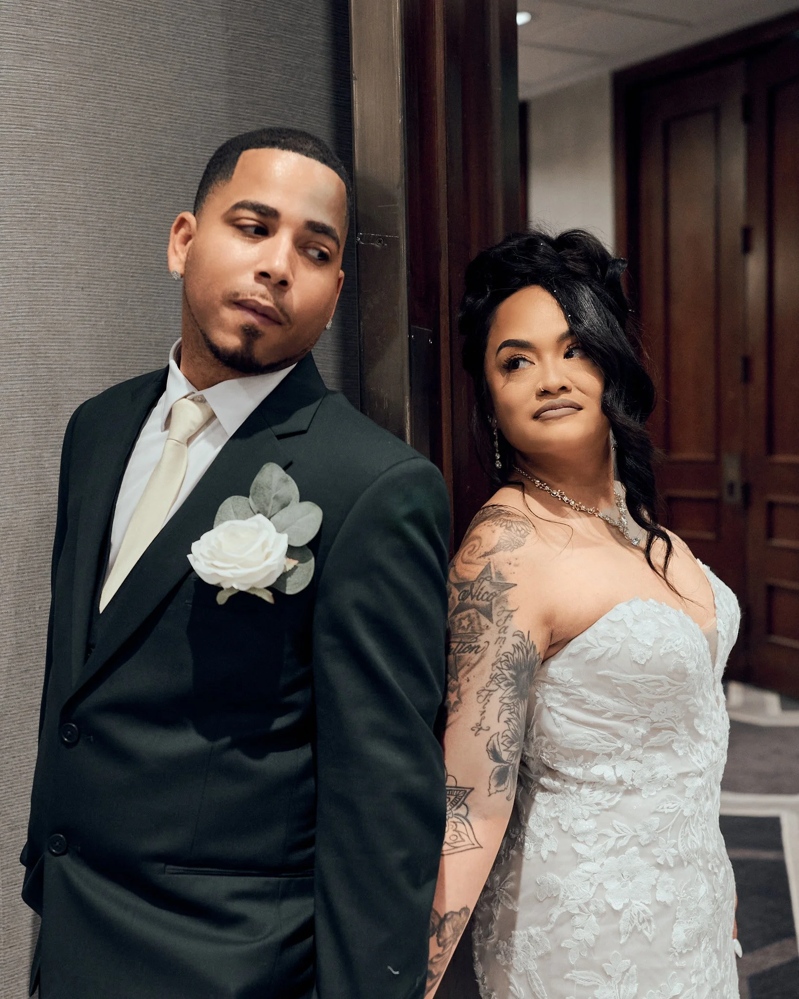 A groom and bride standing back to back, leaning against a wall, at a formal wedding. The groom is wearing a black suit with a white shirt and a white boutonniere. The bride is wearing a white strapless gown with floral lace details and has tattoos o
