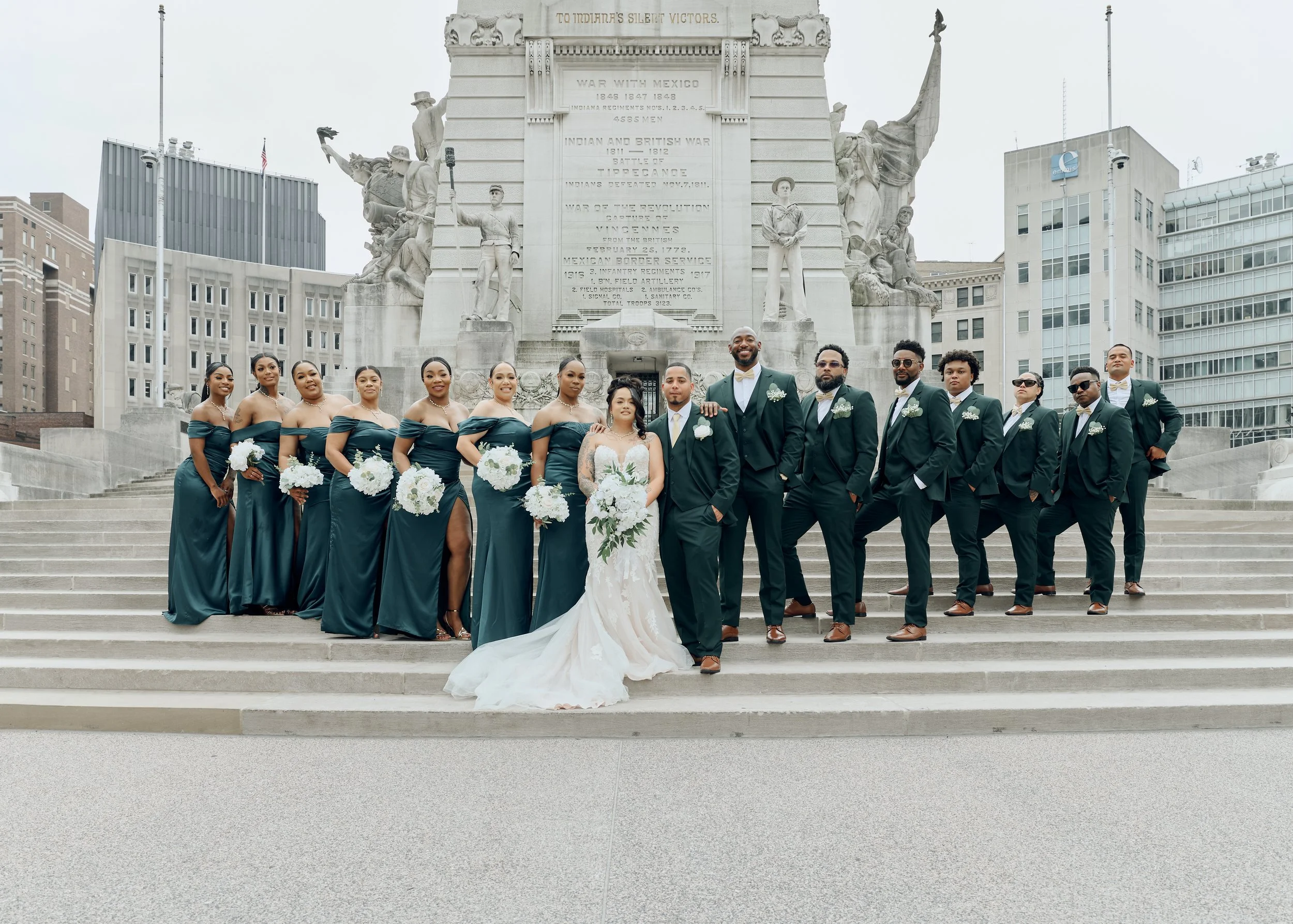 A wedding party standing on steps in front of a large monument with statues and engraved text, with city buildings in the background. The group includes the bride in a white wedding dress, groom in a dark tuxedo, and bridesmaids and groomsmen dressed