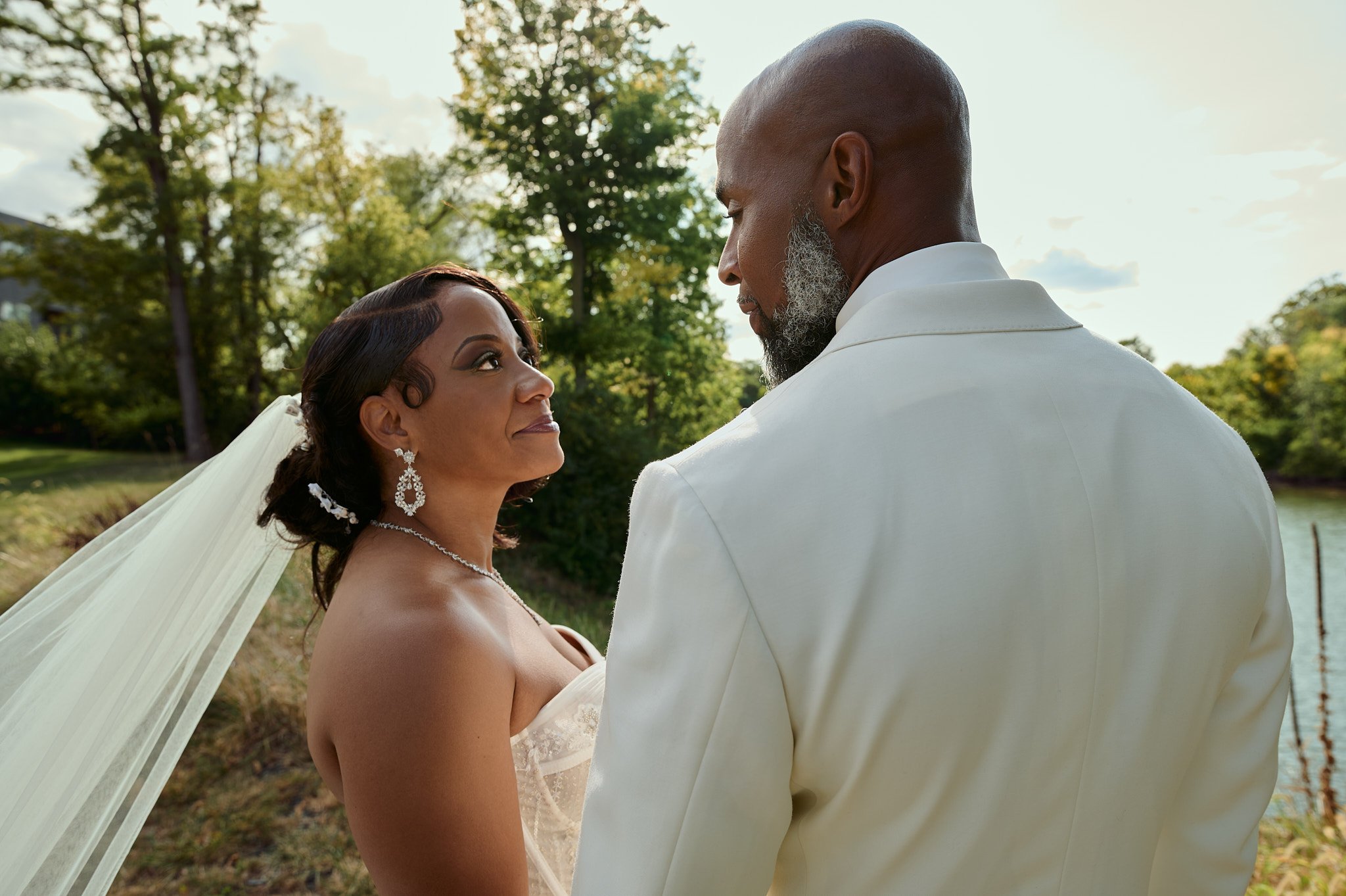 A bride and groom face each other outdoors, with trees and a body of water in the background, during a wedding ceremony.