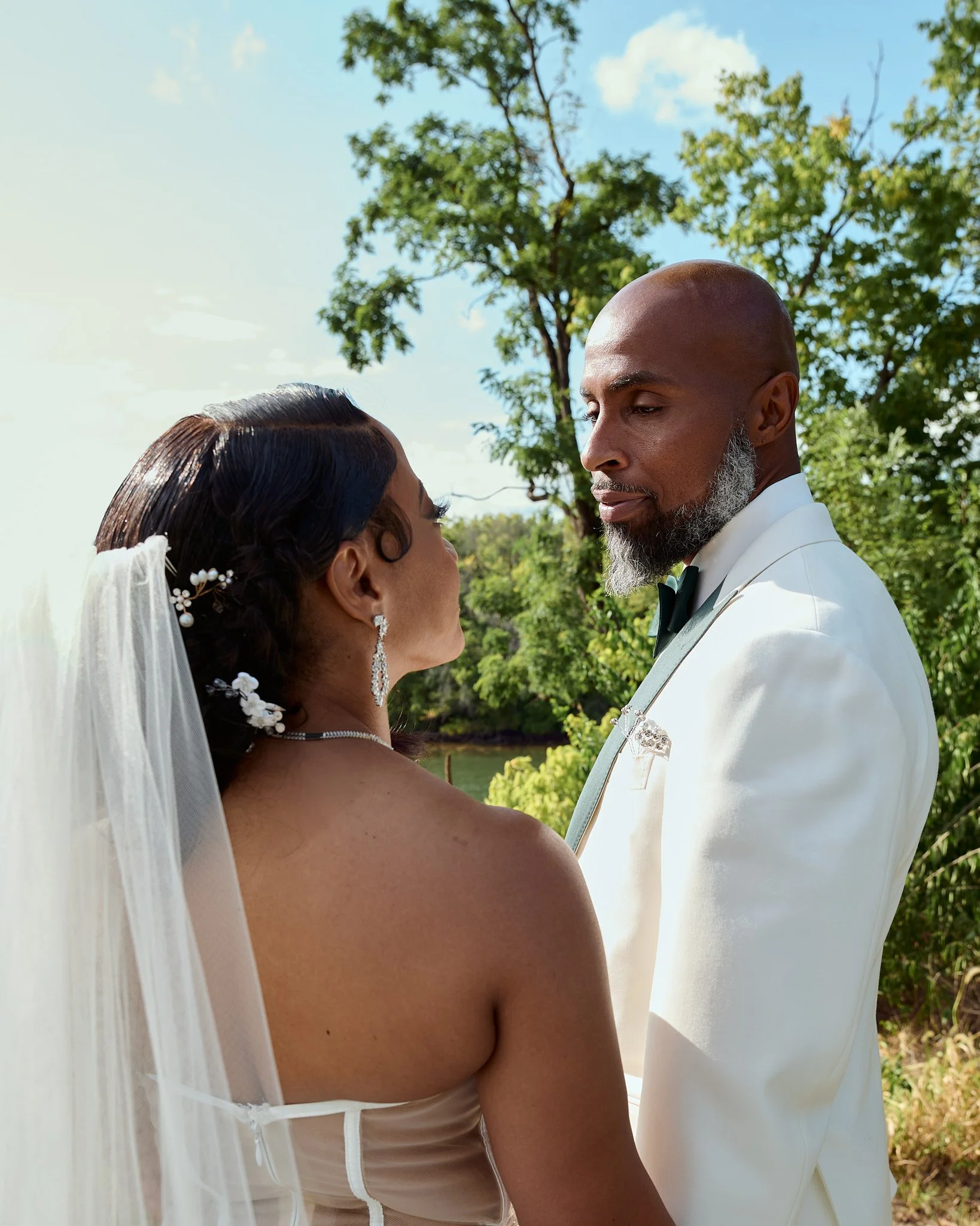 A bride and groom sharing an intimate moment outdoors, with greenery and a tree in the background under a partly cloudy sky.