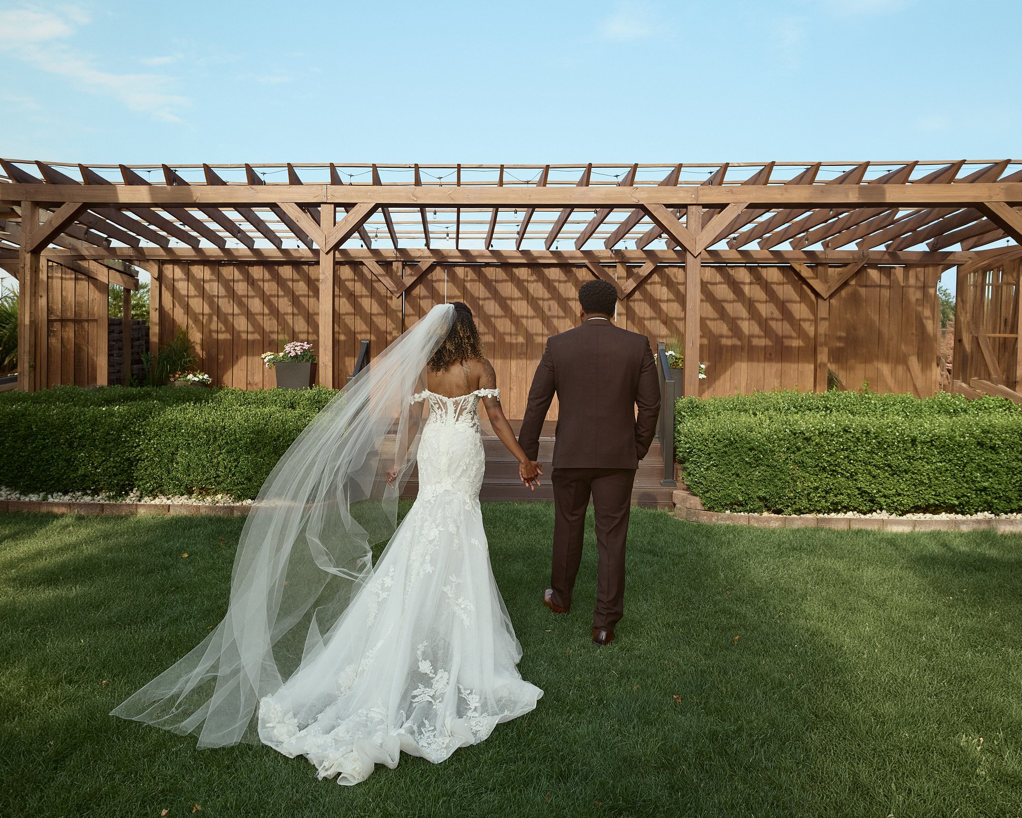 A wedding couple holding hands, walking away from the camera, towards a wooden altar structure outdoors under a blue sky.