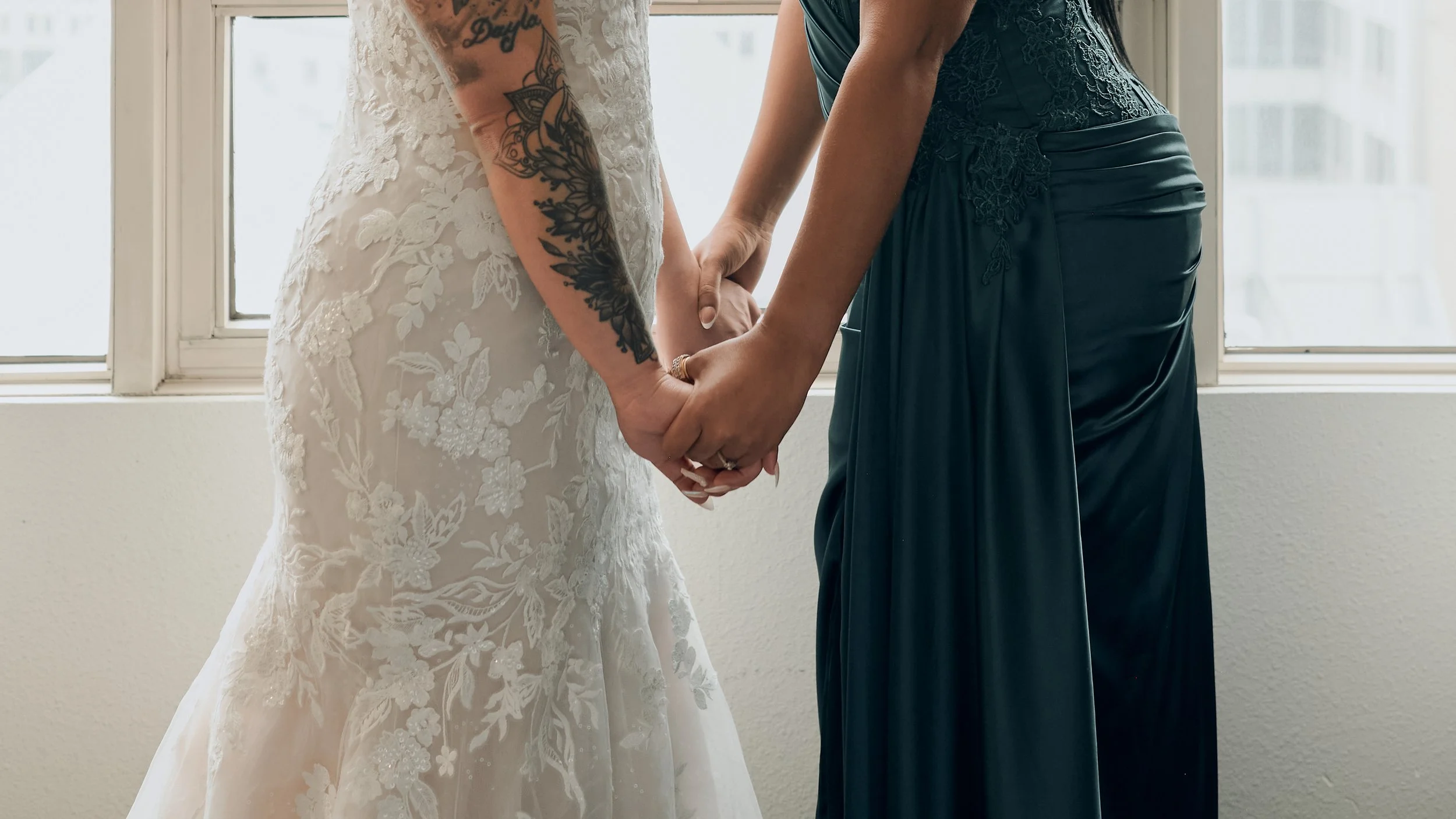 Two women holding hands, dressed in wedding attire, standing in front of a window.