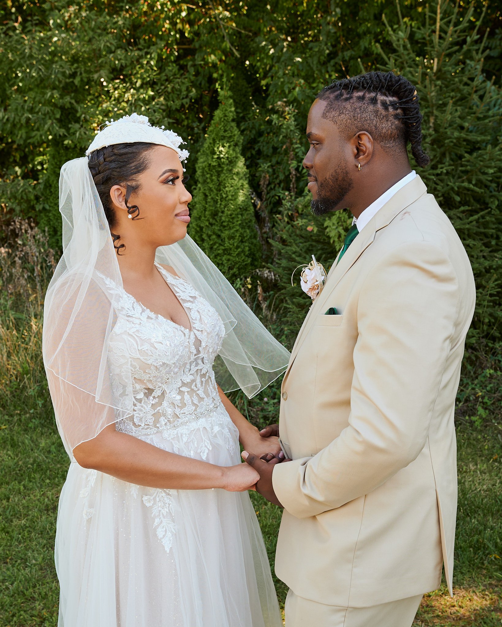 A bride and groom holding hands outdoors, facing each other, with green trees in the background.