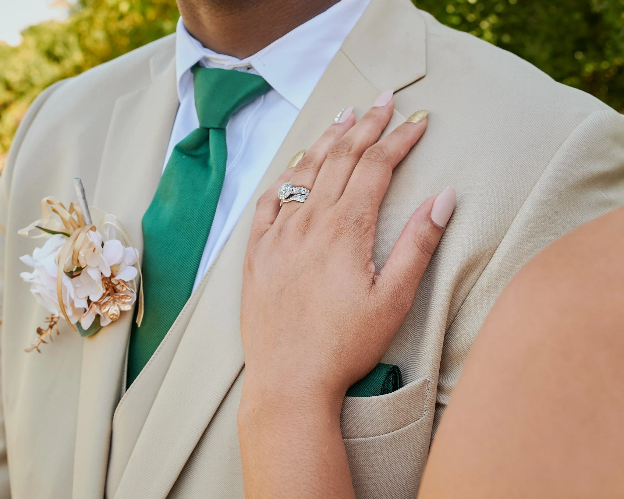 A close-up of a bride's hand with wedding rings resting on a groom's chest, who is wearing a light beige suit, white shirt, and green tie, with a small flower boutonniere on his lapel.