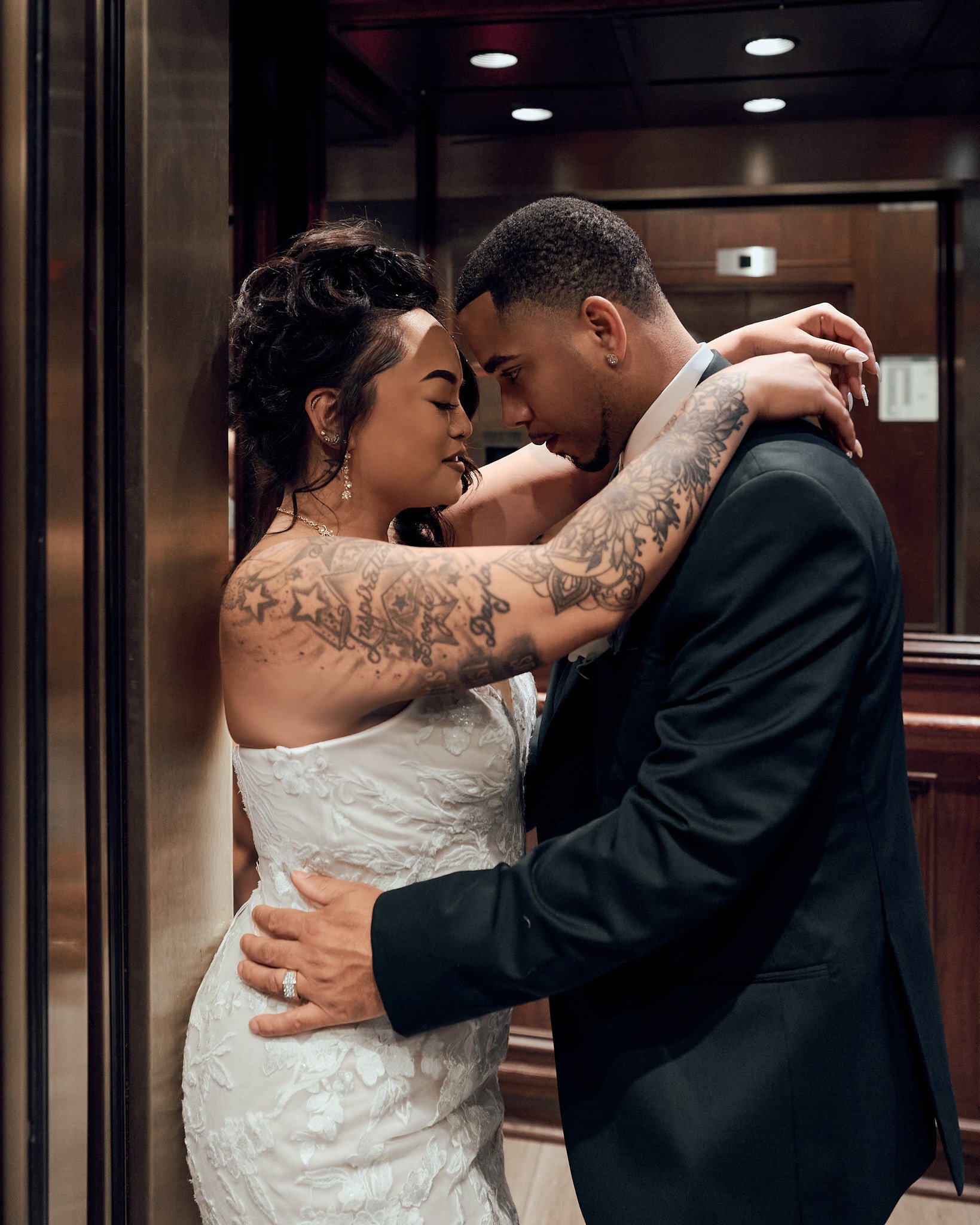 A couple dressed in wedding attire sharing an intimate moment in an elevator, with their foreheads touching and eyes closed.
