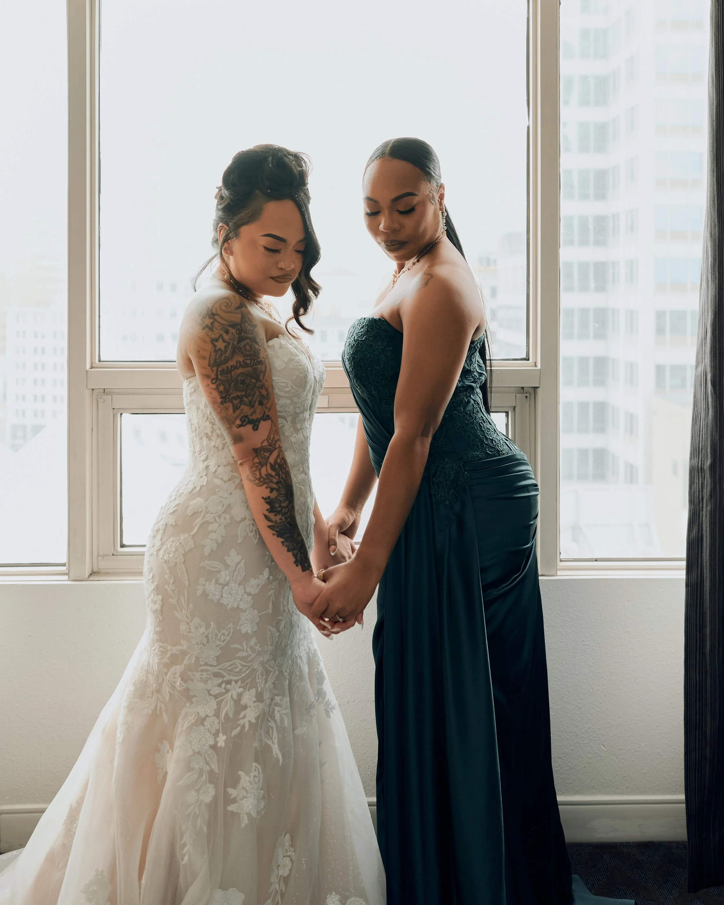 Two women holding hands and standing close together in front of a large window, one in a wedding dress and the other in a formal dress.