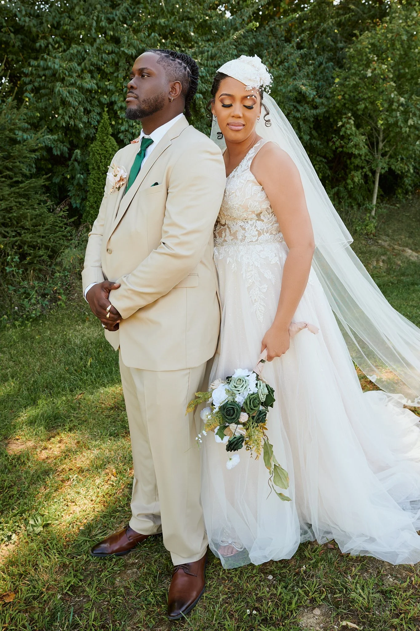 A bride and groom stand close together outdoors, the bride holding a bouquet of white and green flowers, both dressed in wedding attire with a grassy and wooded background.