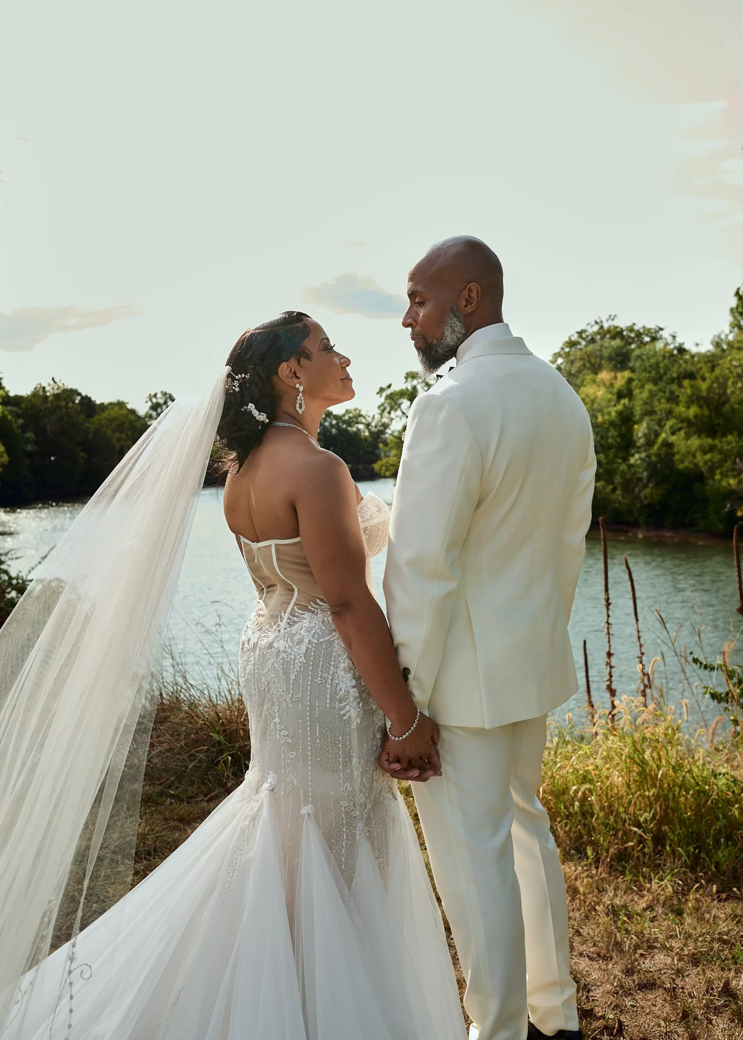 A bride and groom holding hands by a river with trees in the background on their wedding day.