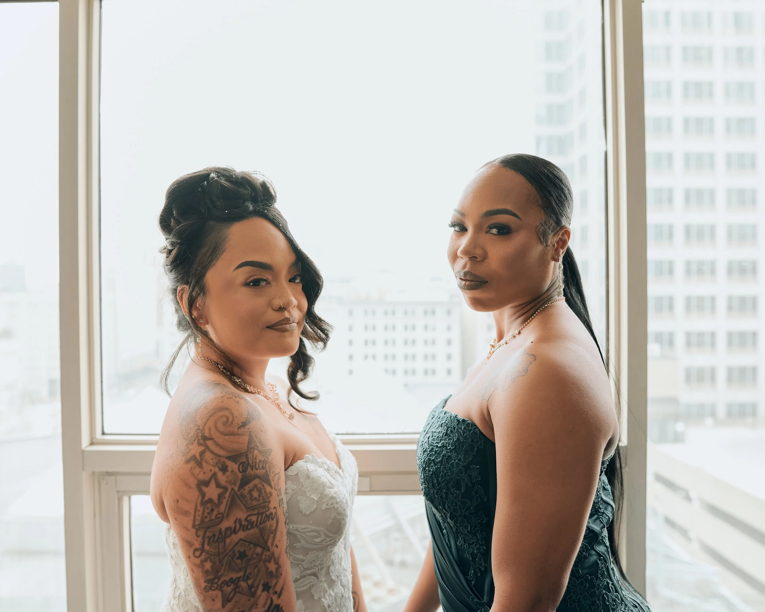 Two women standing by a large window with city buildings in the background, dressed in elegant outfits with tattoos visible on one woman's arm, both with confident expressions.