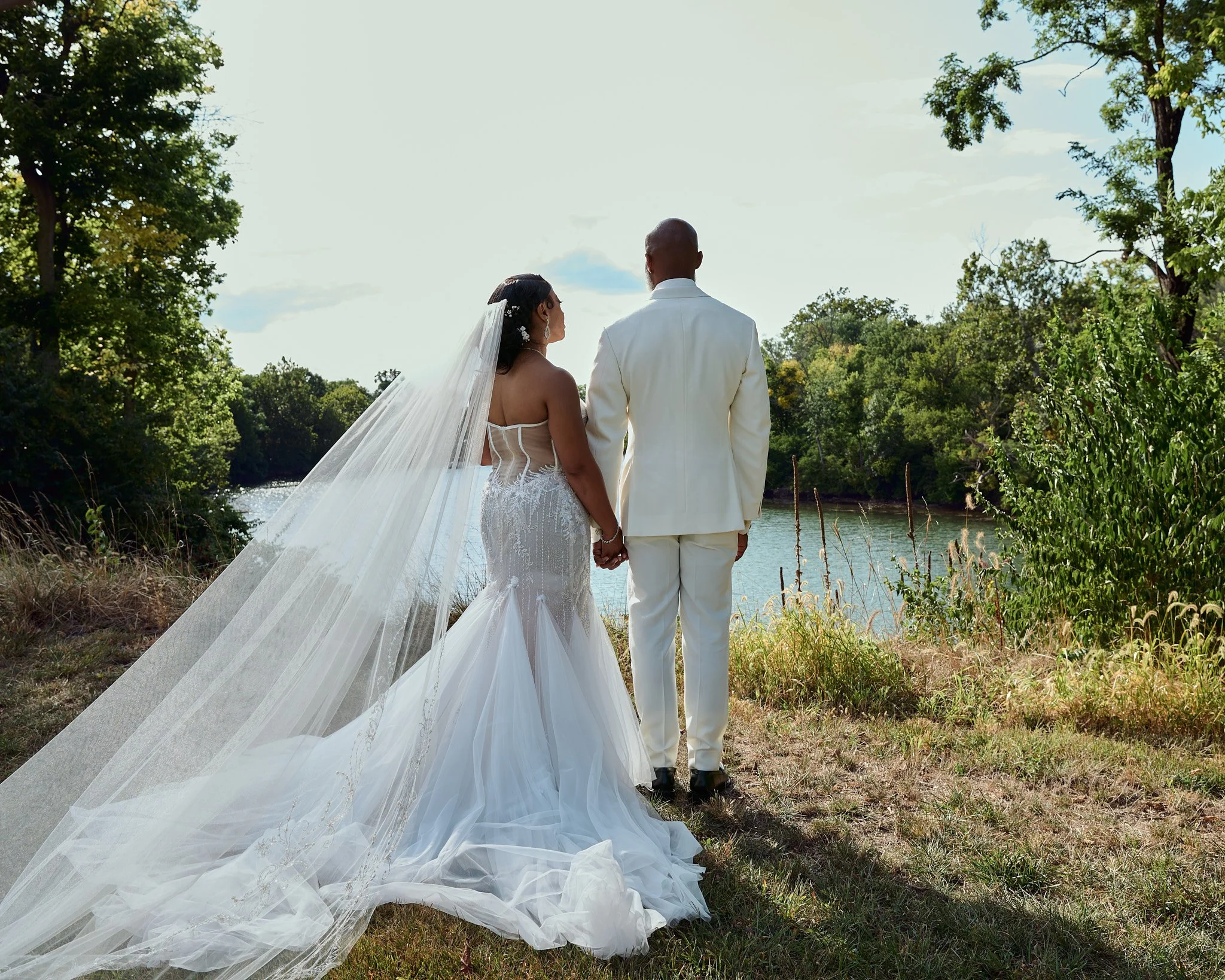 A bride and groom holding hands, standing by a river in a lush, green outdoor setting during daytime, facing away from the camera.