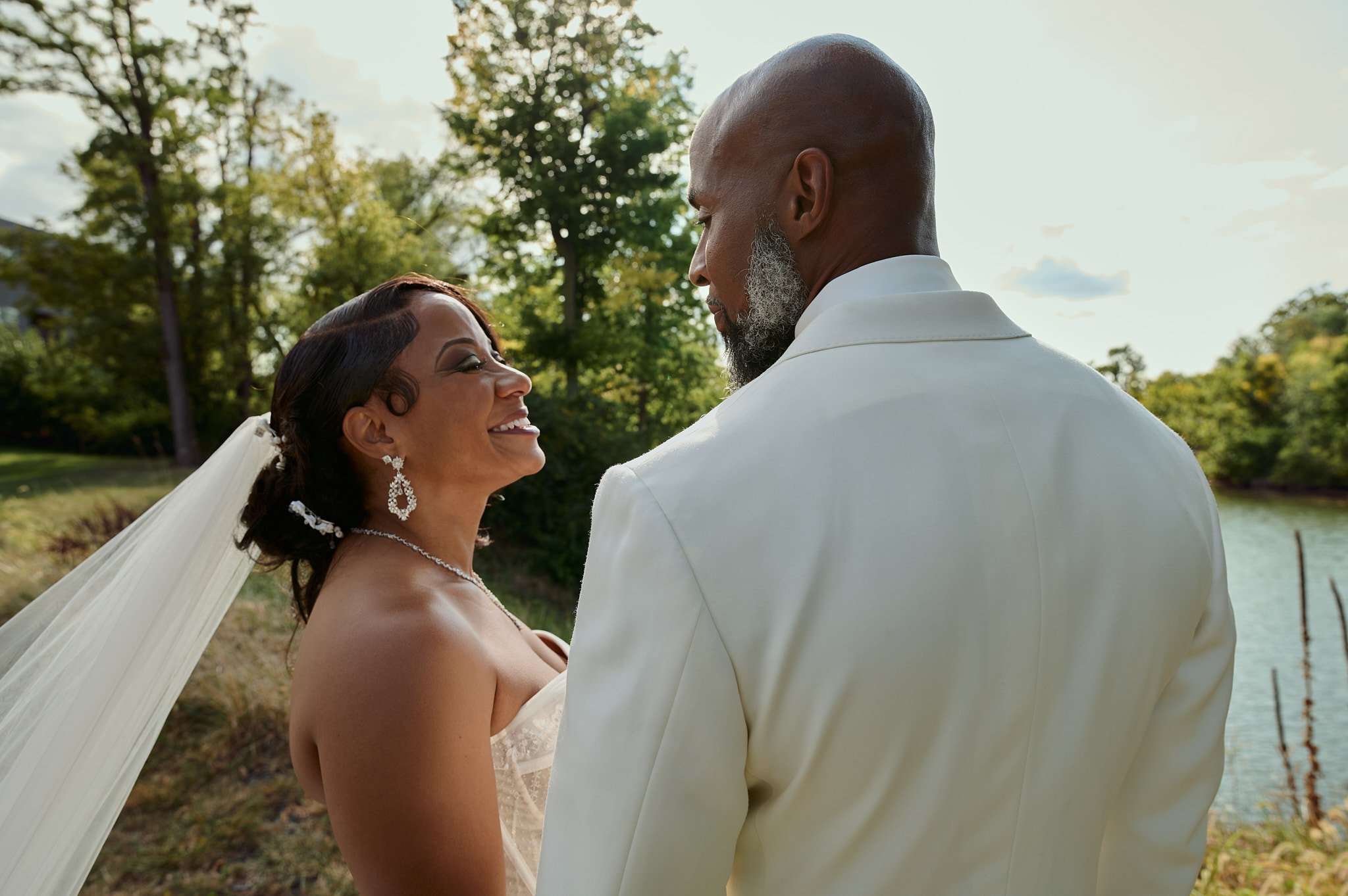 A bride and groom smiling and looking at each other during their outdoor wedding near a river, with trees and a partly cloudy sky in the background.