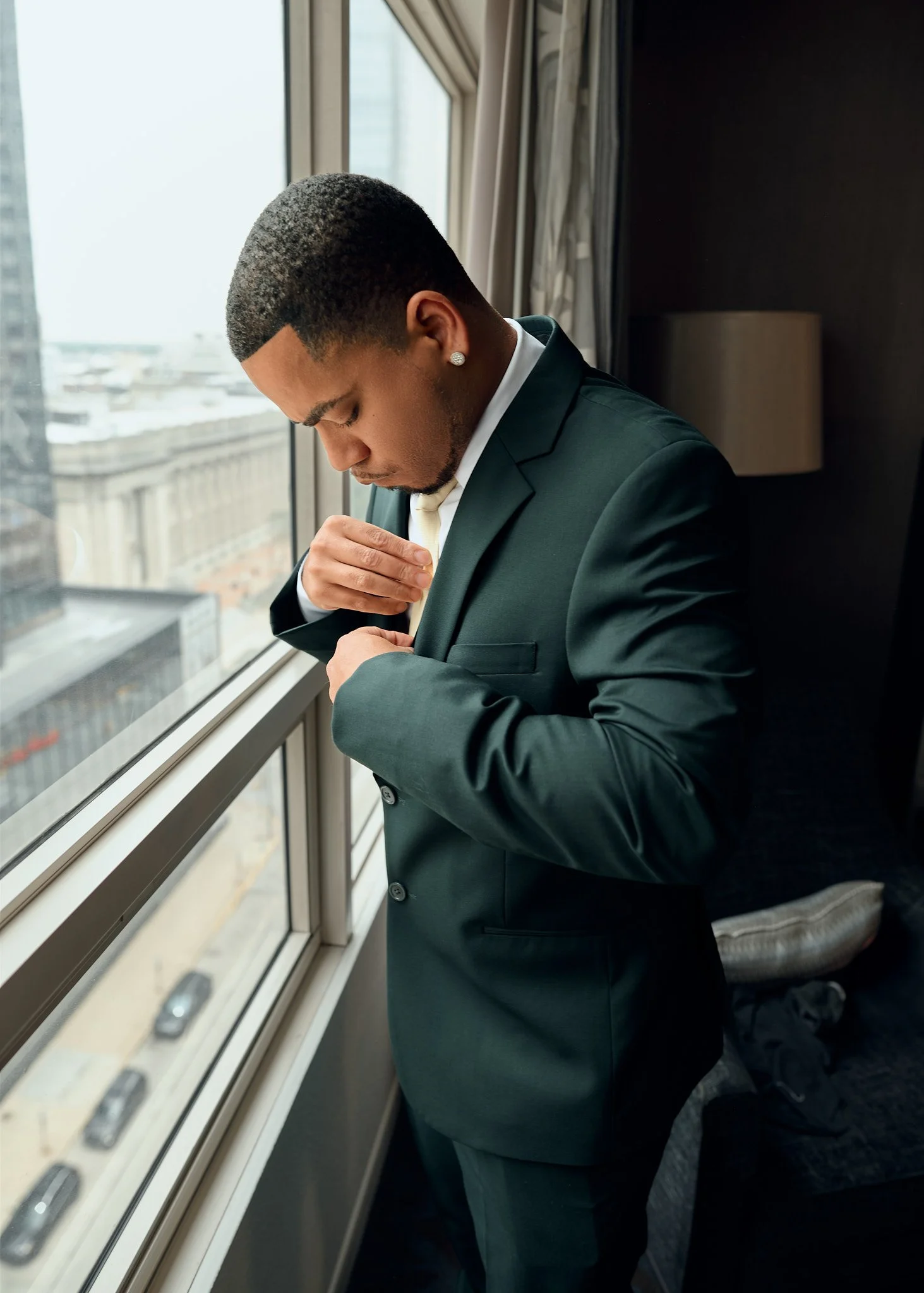 A young man in a dark suit adjusting his jacket while standing by a window in a high-rise hotel room, with city buildings visible outside.