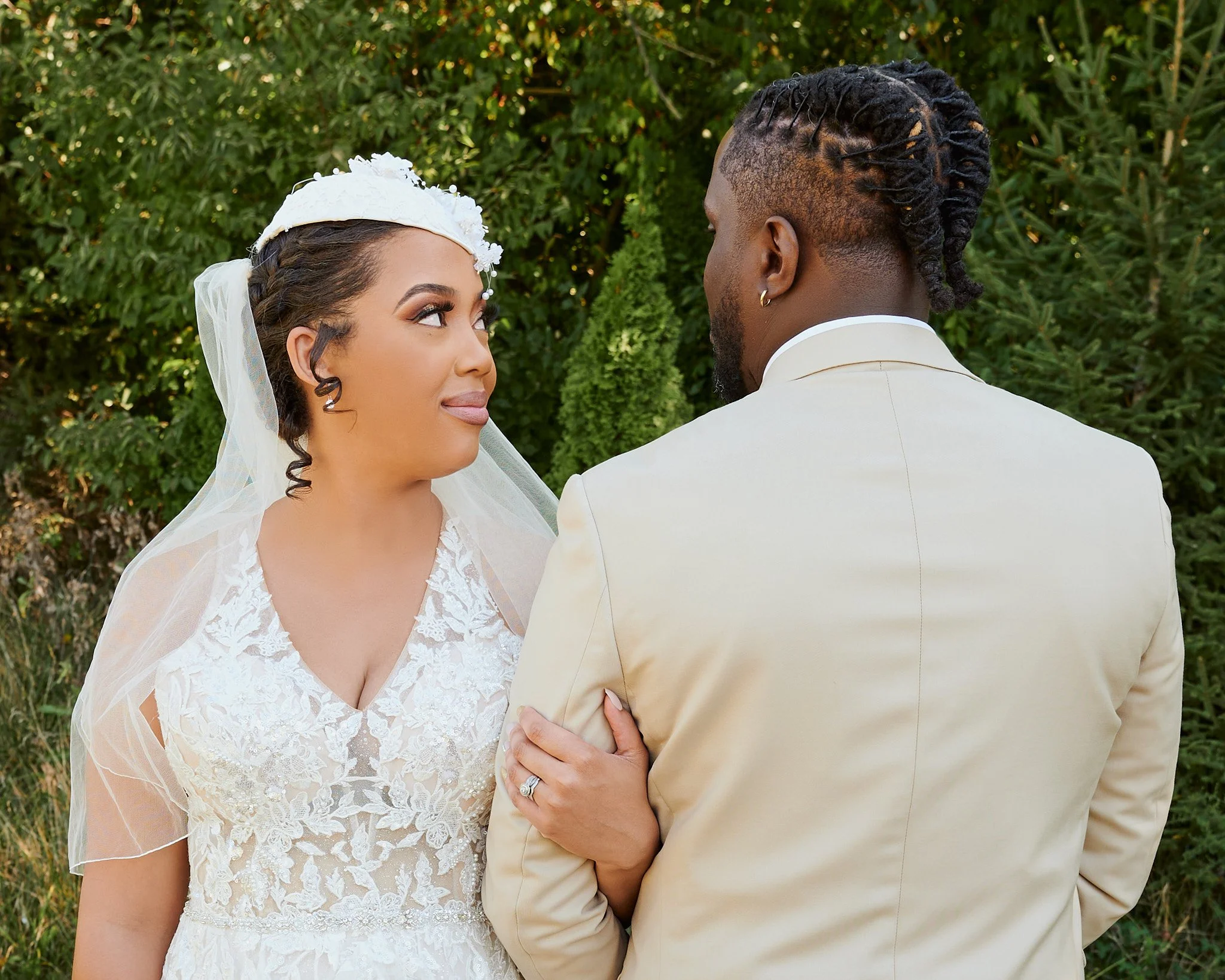A bride and groom standing outdoors, facing each other. The bride is wearing a white lace wedding dress and veil, and the groom is wearing a cream-colored suit. They are holding hands, with the bride's ring visible on her finger, and looking into eac