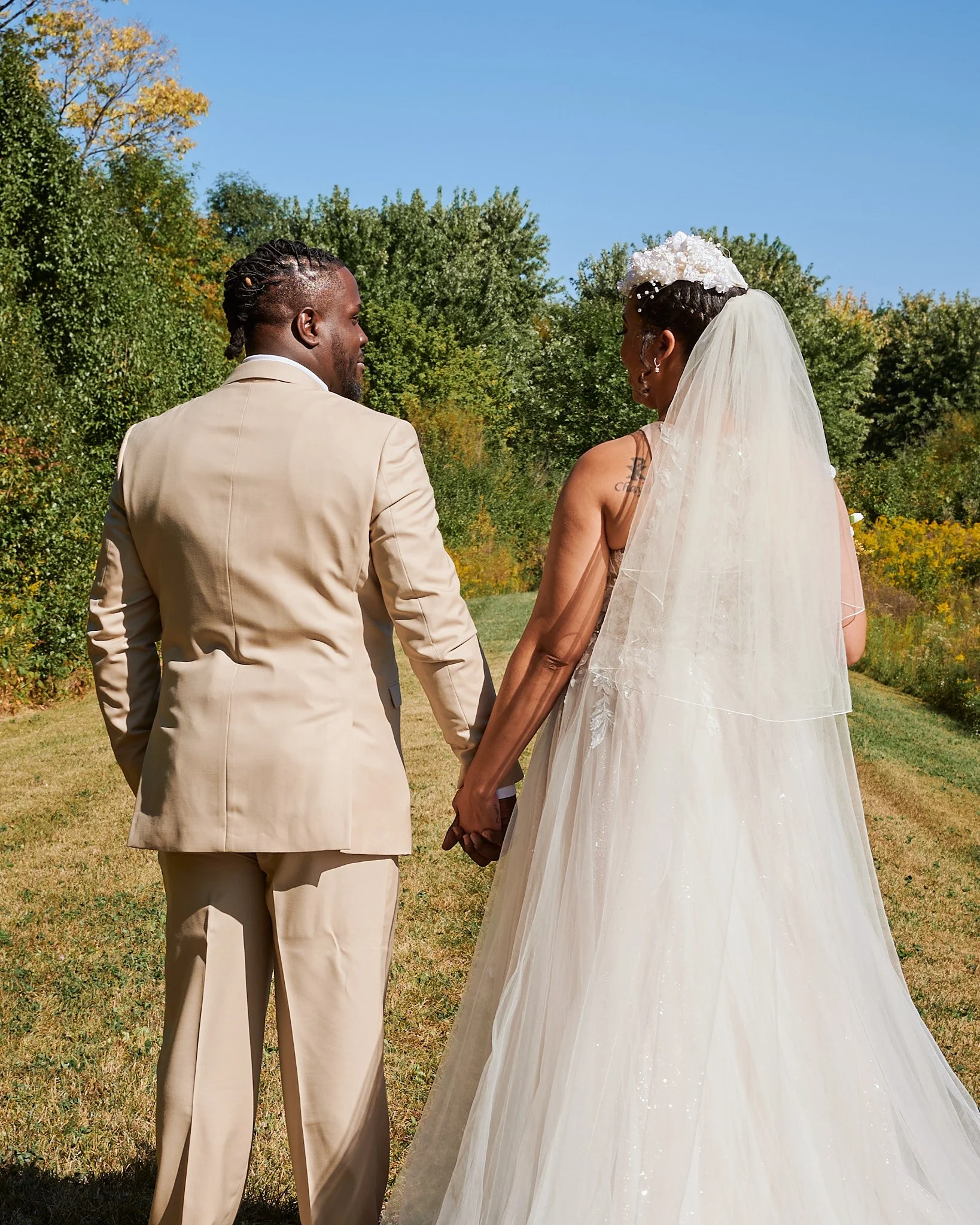 A couple in wedding attire holding hands outdoors on a sunny day, surrounded by green trees and a grassy area.
