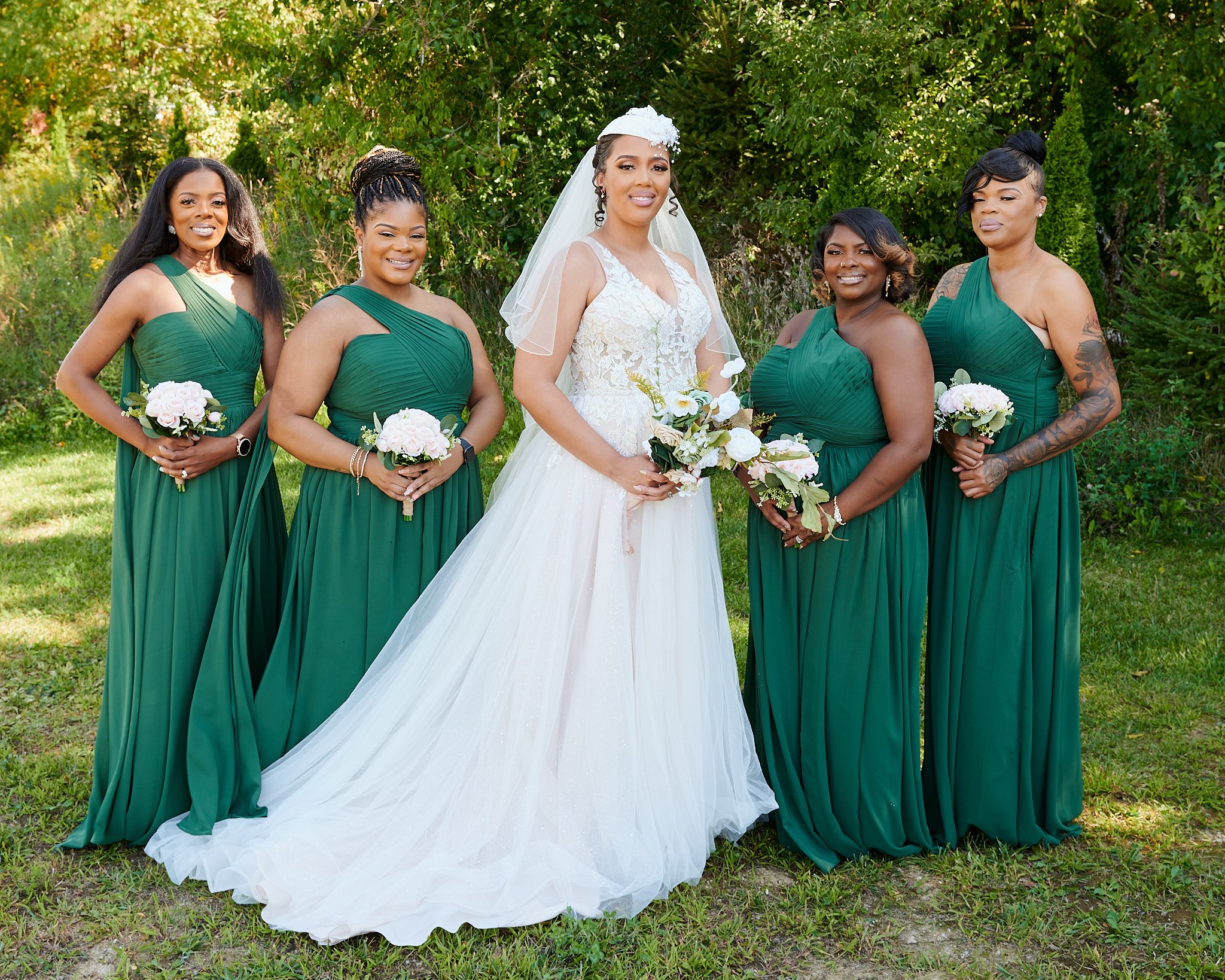 Bride in white wedding dress standing with five bridesmaids in green dresses outdoors with trees in the background.