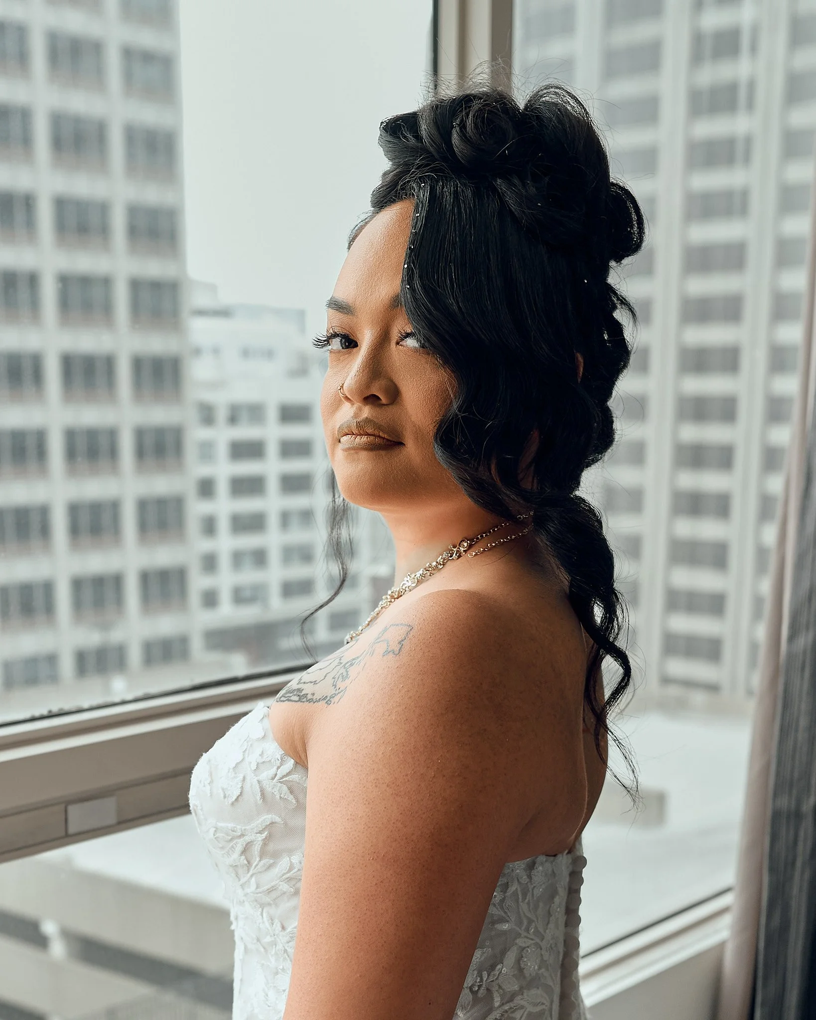 A woman with dark, styled hair, wearing a white strapless dress, standing near a large window with a cityscape of high-rise buildings in the background.