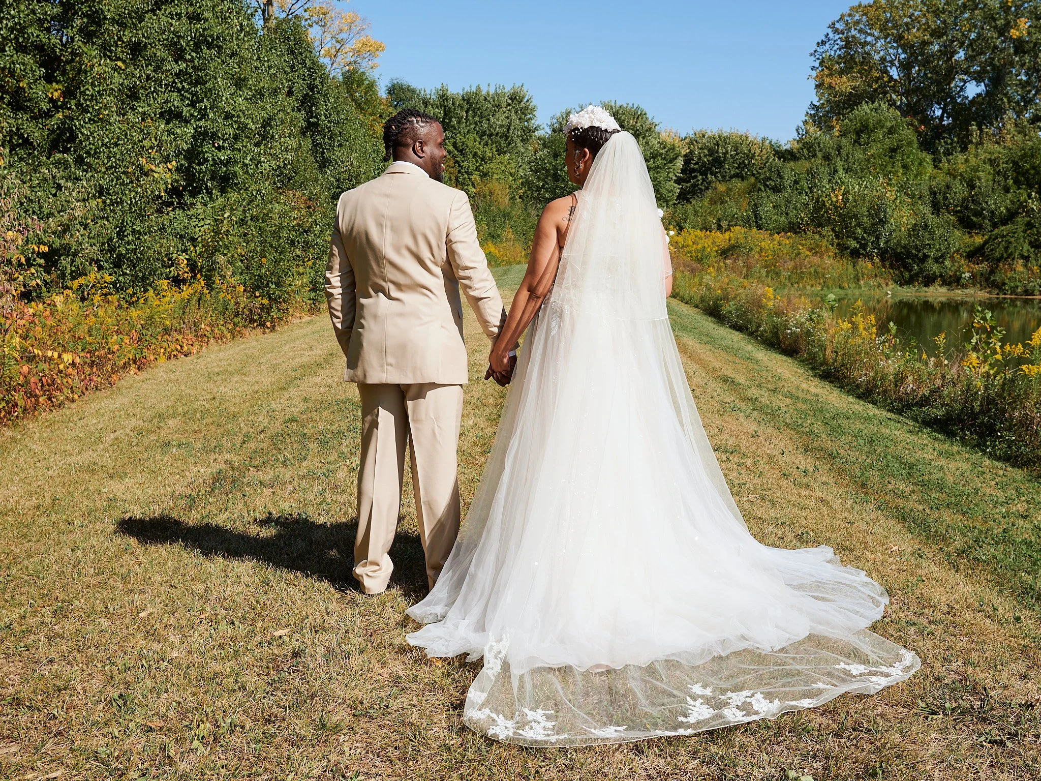 A bride and groom holding hands and walking in a grassy outdoor area, surrounded by trees with fall foliage, near a small body of water, on a sunny day.