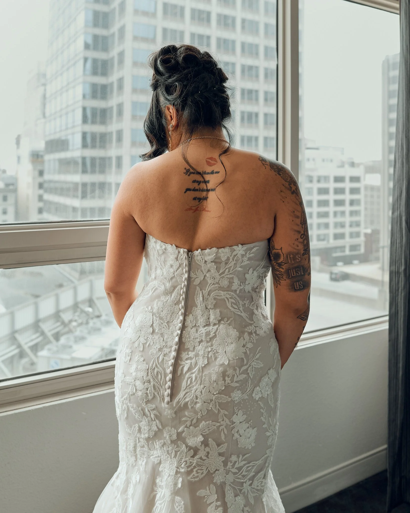 Woman in a strapless white wedding dress with lace details, standing by a large window overlooking a cityscape of tall buildings during daytime.