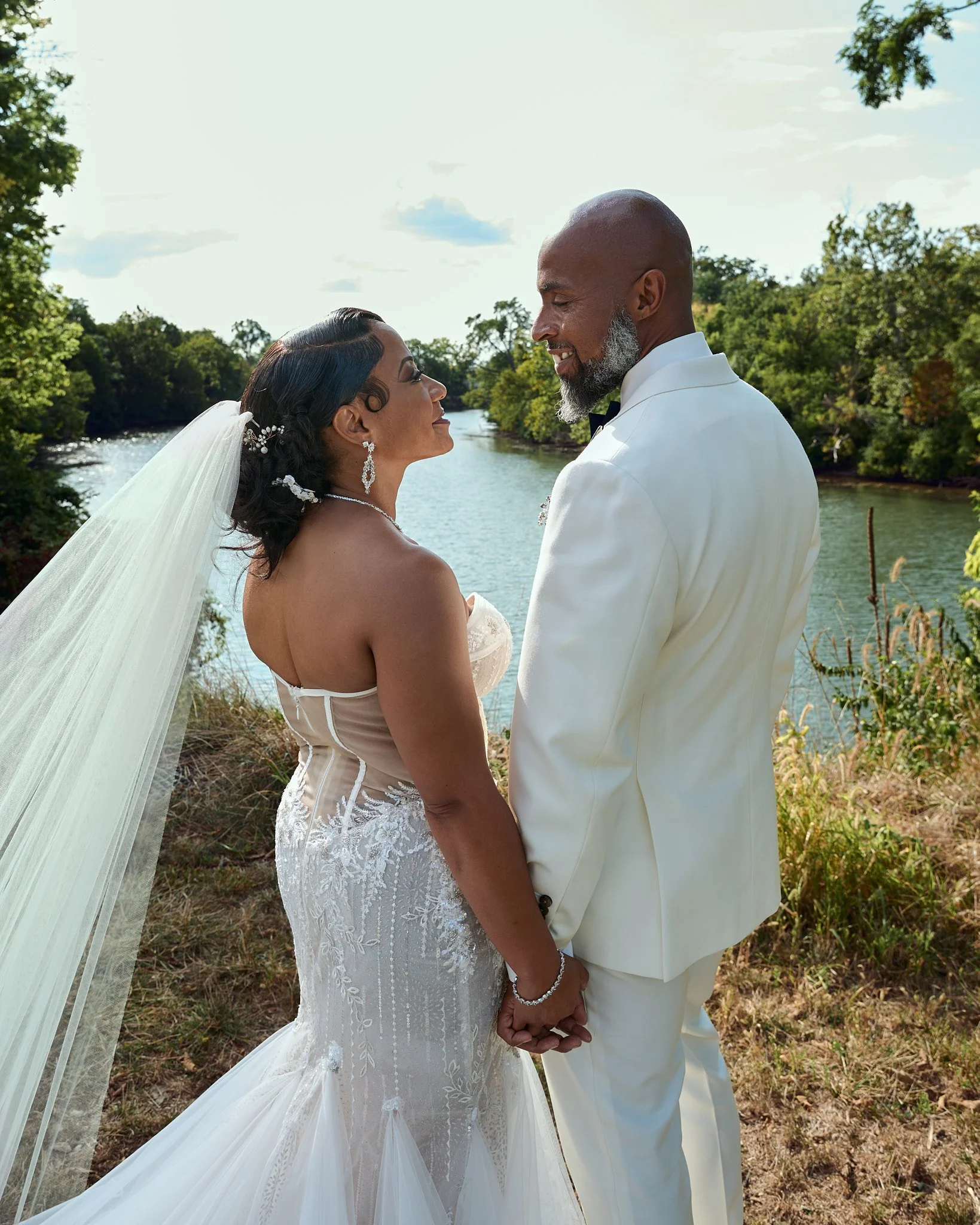 A bride and groom holding hands, facing each other outdoors near a river, with trees in the background on a sunny day.