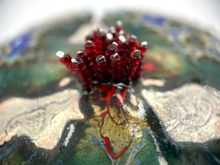 detail of round handmade enamel brooch with blue and green and silver organic pattern and red beads