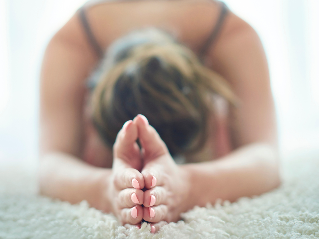 Nahaufnahme einer Person beim Yoga im Kind-Position auf einem weichen, hellen Teppich. Die Hände sind in Gebetshaltung zusammengeführt.