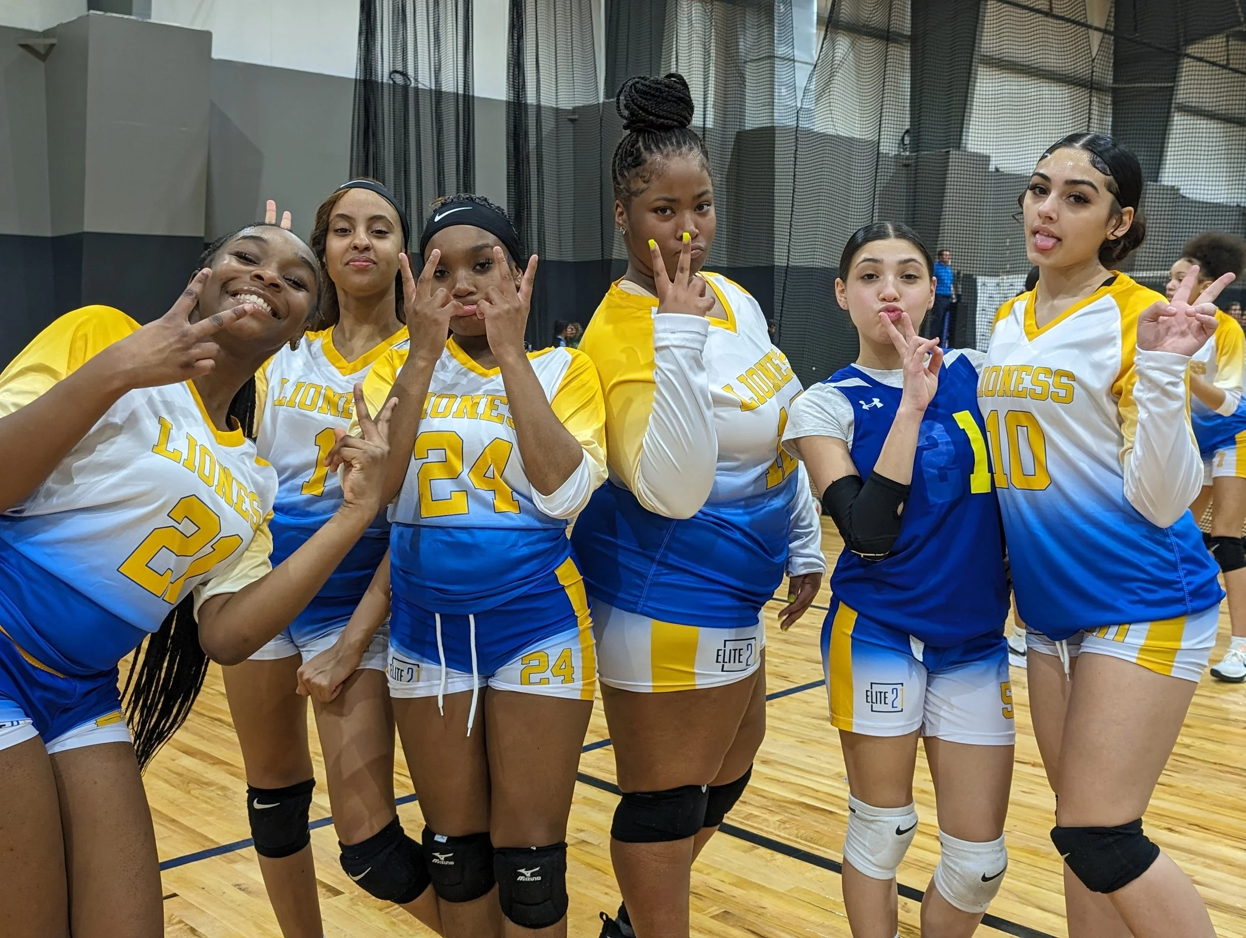 A group of six young women volleyball players wearing yellow, blue, and white uniforms posing together on an indoor volleyball court.