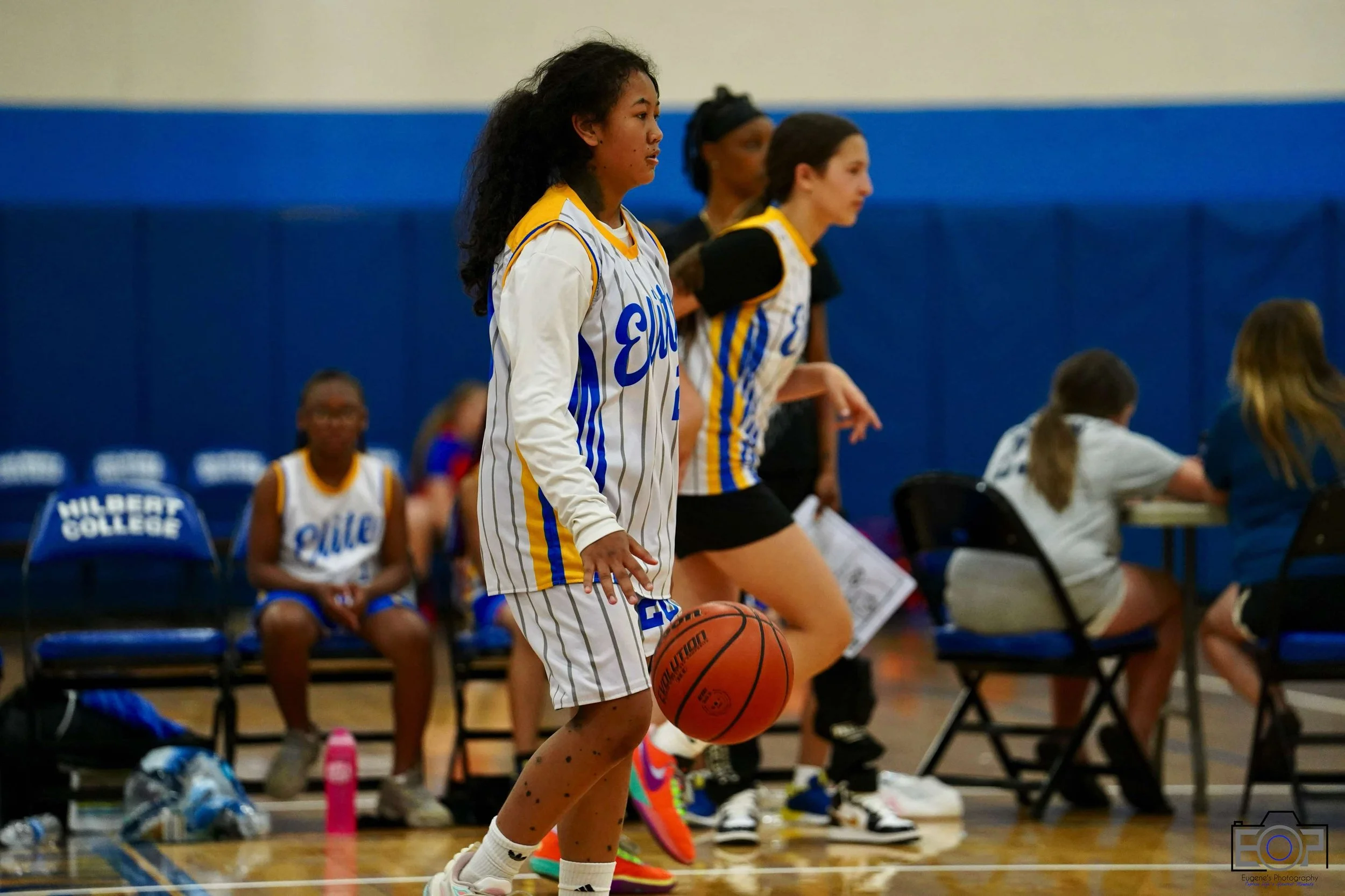 Young girl playing basketball during a game, with teammates and spectators in the background at Wilbert College gym.