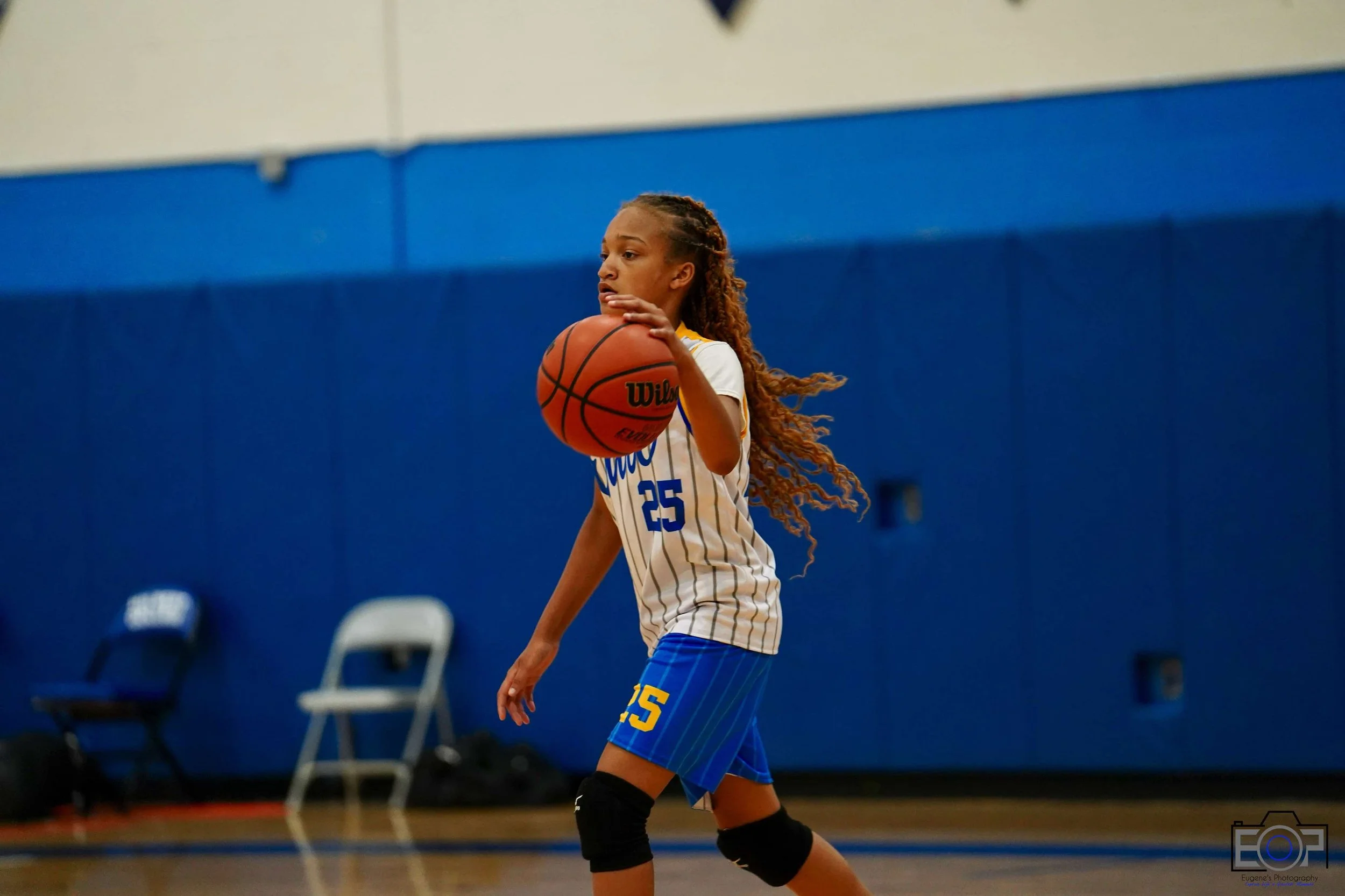 Young girl with long curly hair playing basketball indoors, wearing a white jersey with blue and yellow accents, number 25, black knee pads, and holding a basketball.