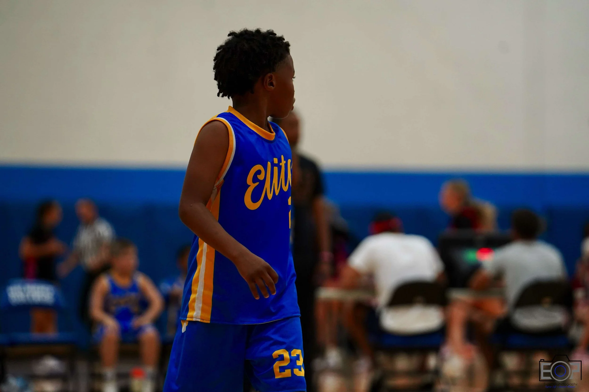 A young basketball player wearing a blue and yellow uniform with the number 23 on it, standing on the court during a game or practice, with other players and spectators visible in the background.