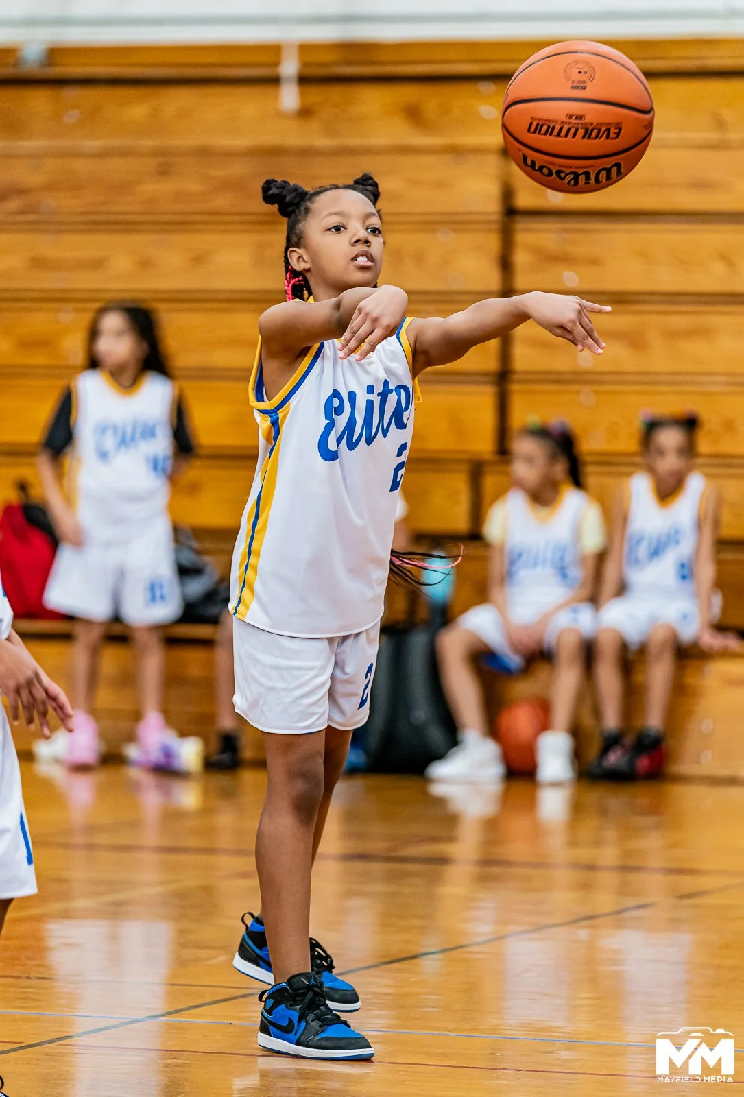 A young boy in a blue t-shirt and shorts dribbling a basketball in a gymnasium with markings on the hardwood floor.