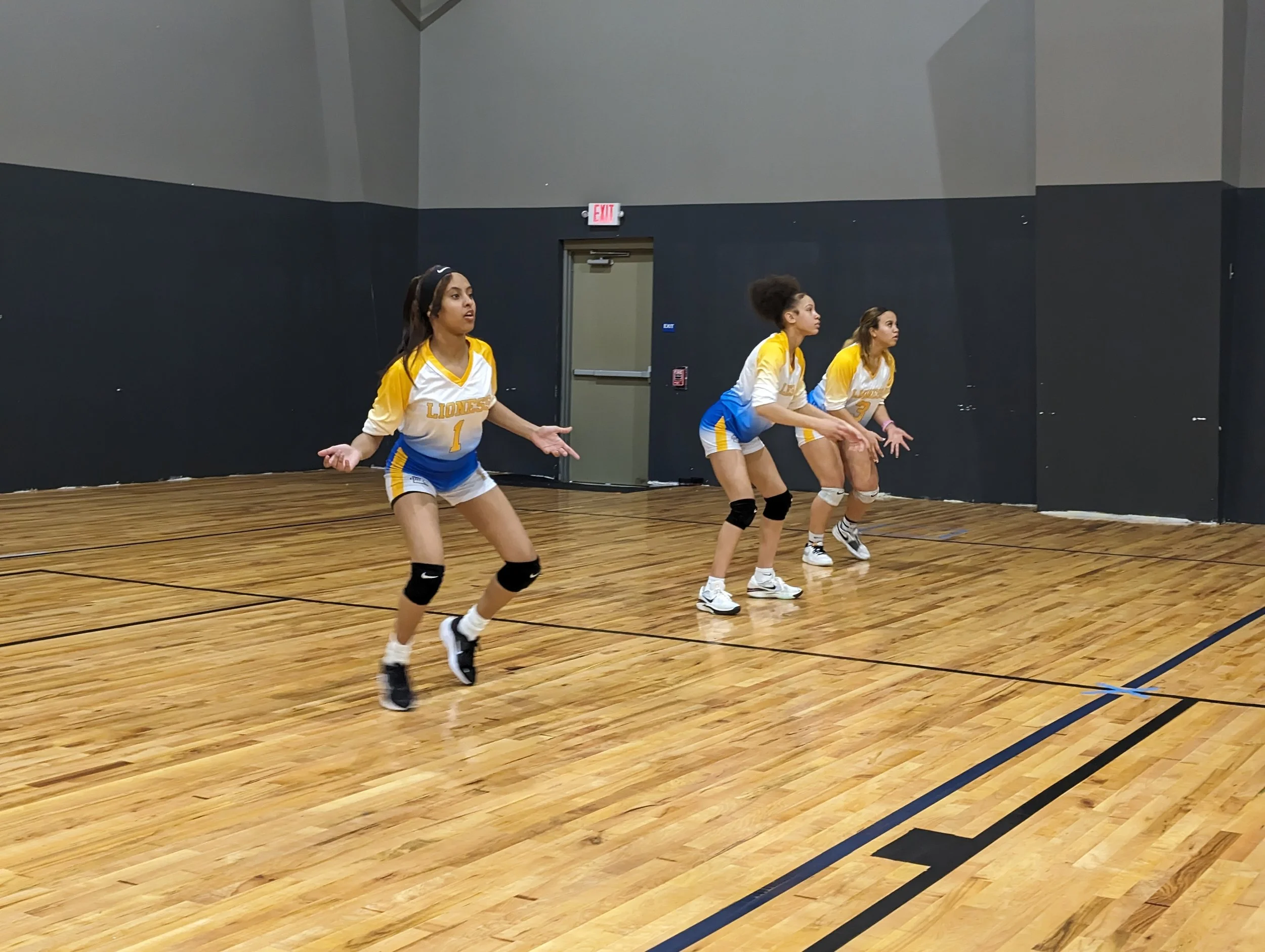 Four female volleyball players in yellow and white uniforms practicing or warming up on an indoor wooden court.
