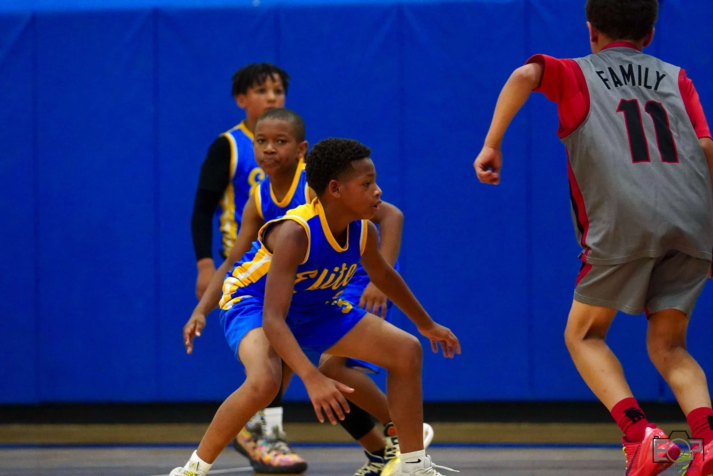 Young boys playing basketball in a gym, with a blue wall in the background. One boy in a gray and red uniform has his back to the camera, while three others in blue and yellow uniforms are facing him, one in a defensive stance.