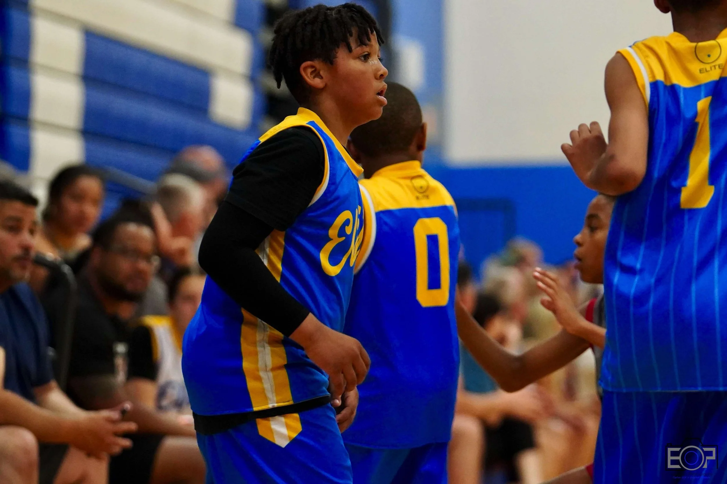 A group of young basketball players practicing on an indoor court, with one player in blue dribbling a basketball.