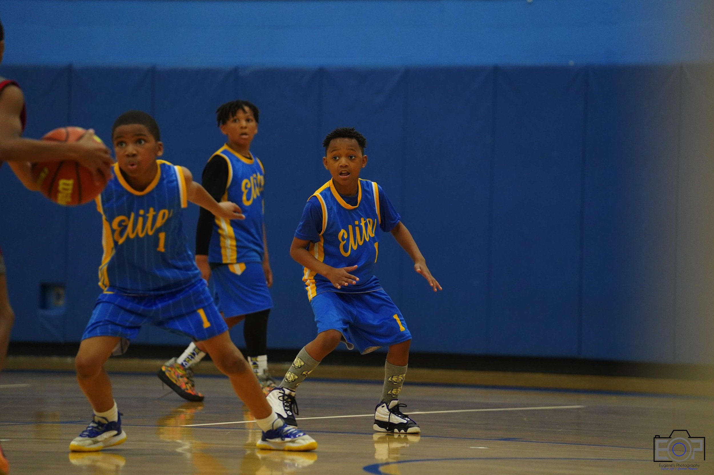 Young boys playing basketball on an indoor court, wearing blue jerseys with yellow letters that spell 'Elite,' and white and black sneakers.