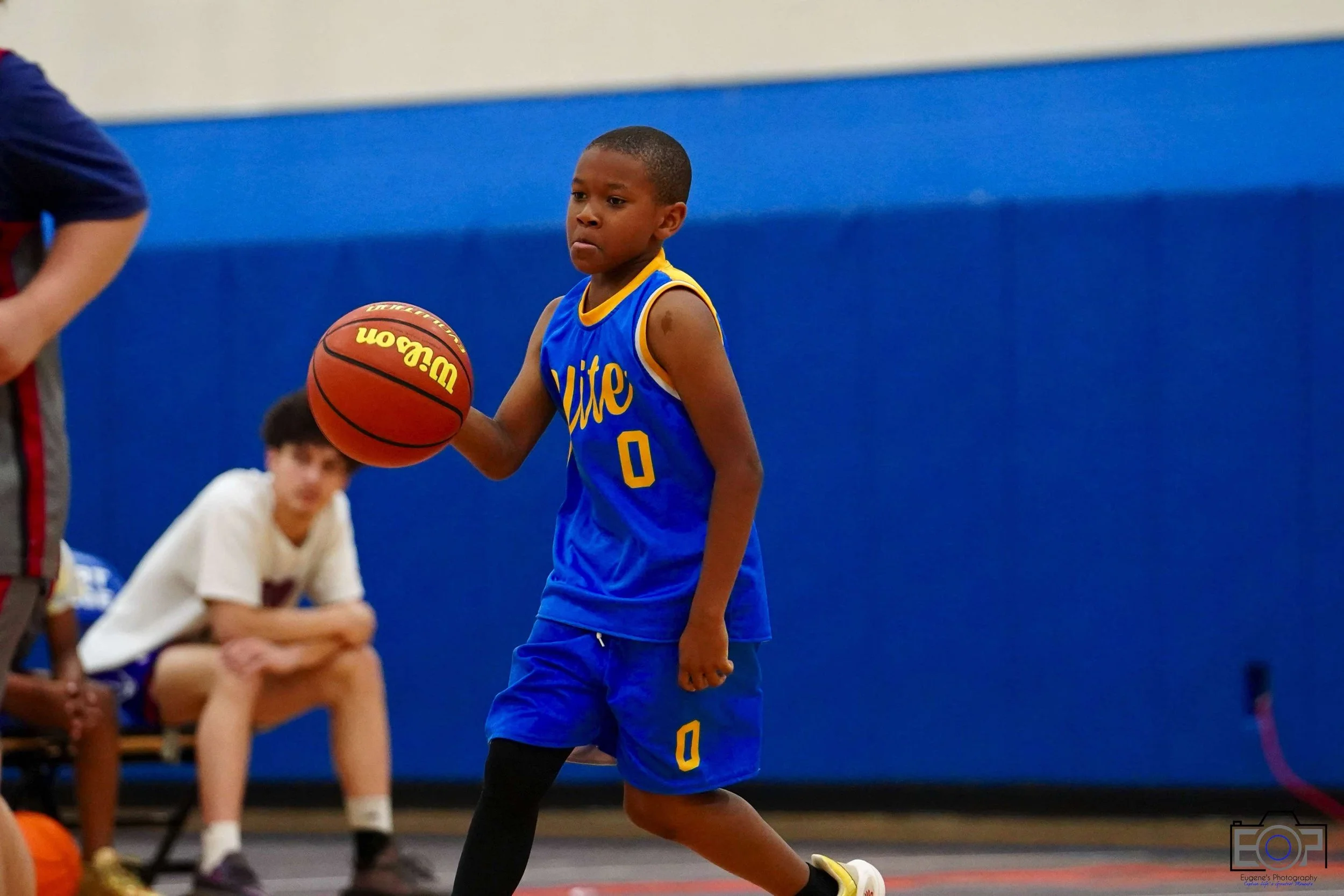 Young boy in blue basketball jersey with yellow trim dribbling a basketball on the court.
