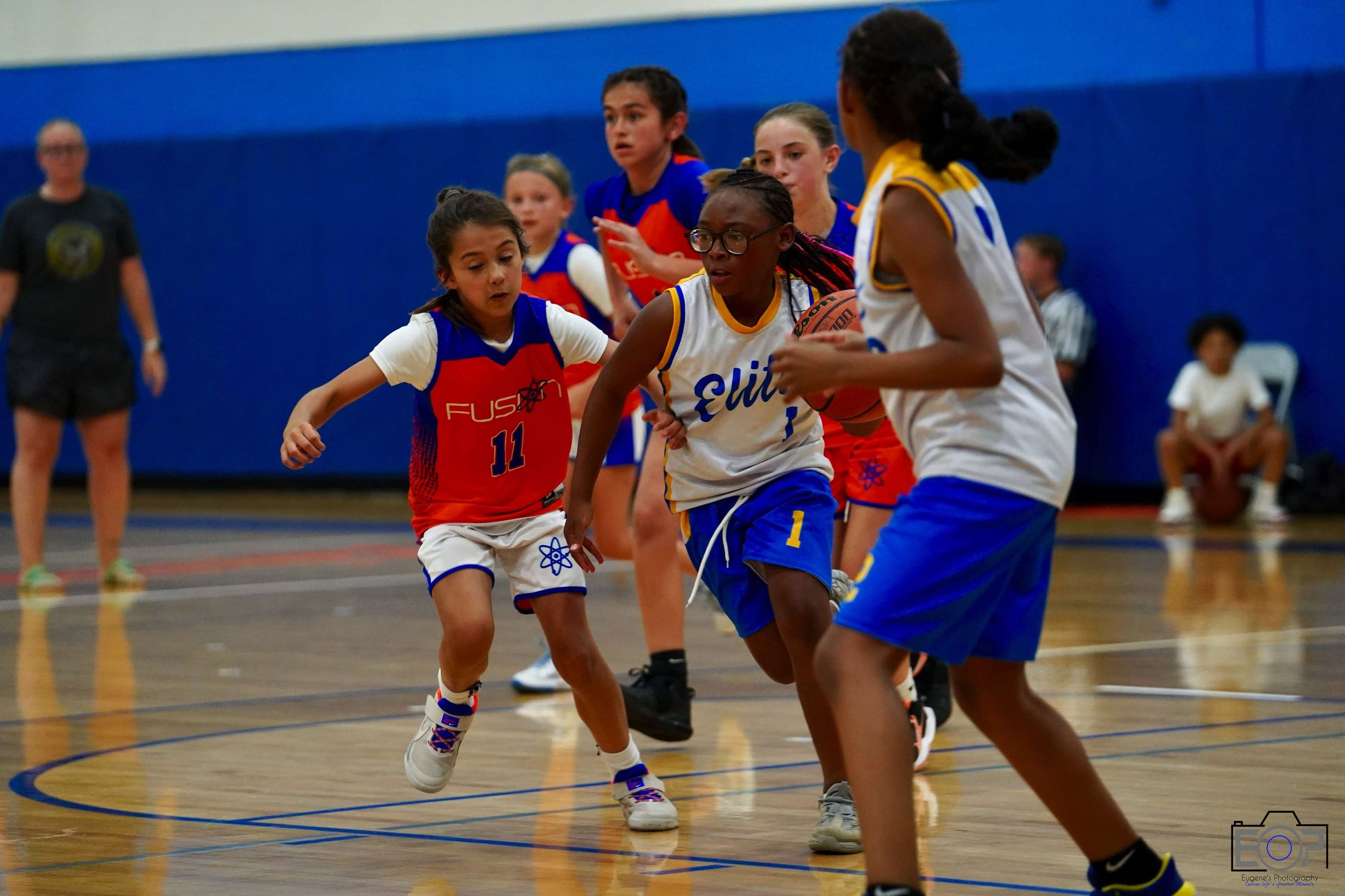 Girls playing youth basketball game in gym with blue walls, wearing uniforms, one girl dribbling and others guarding.