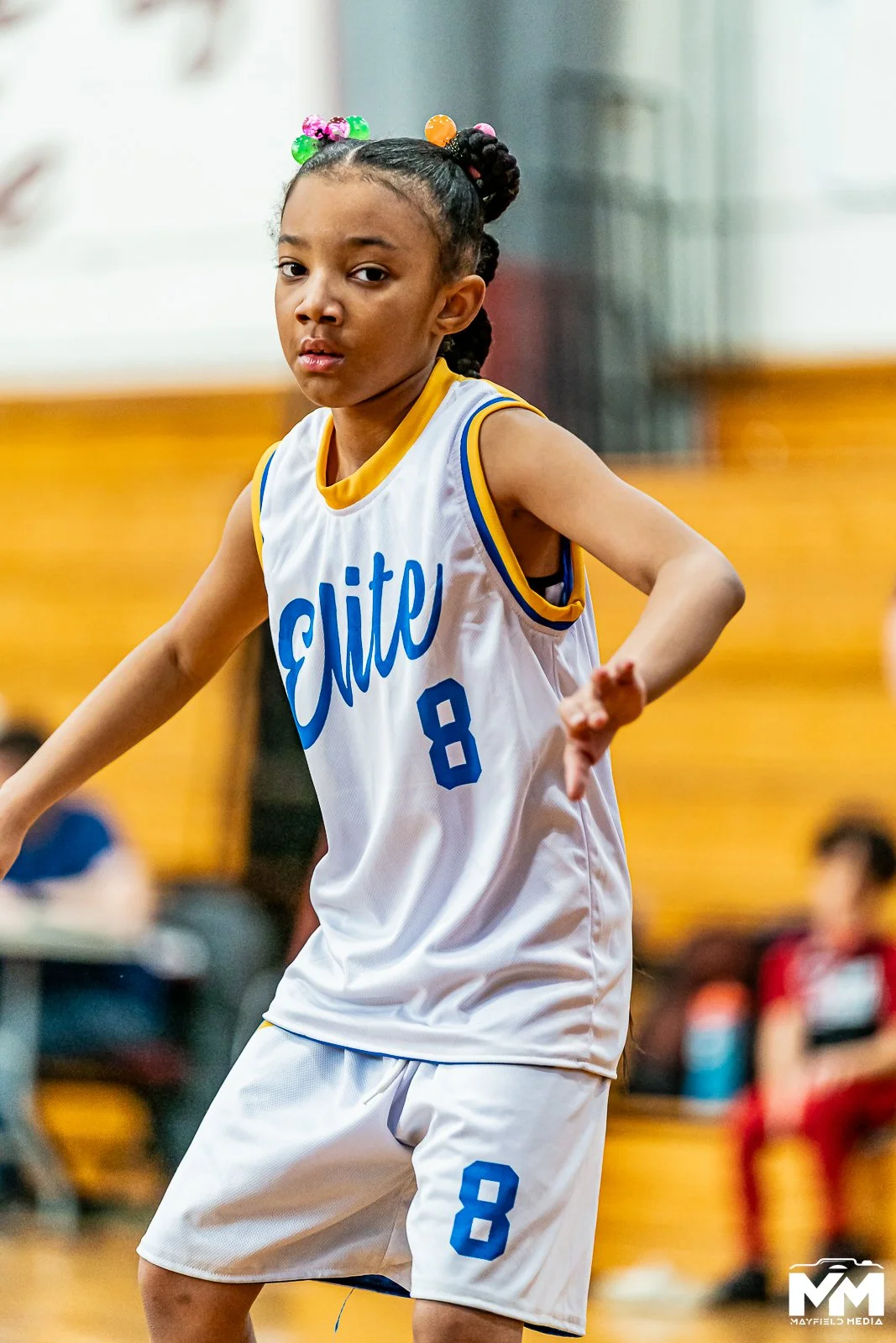 Young girl playing basketball indoors, wearing a white jersey with blue text and the number 8, and purple shorts, with hair styled in braids decorated with colorful beads.