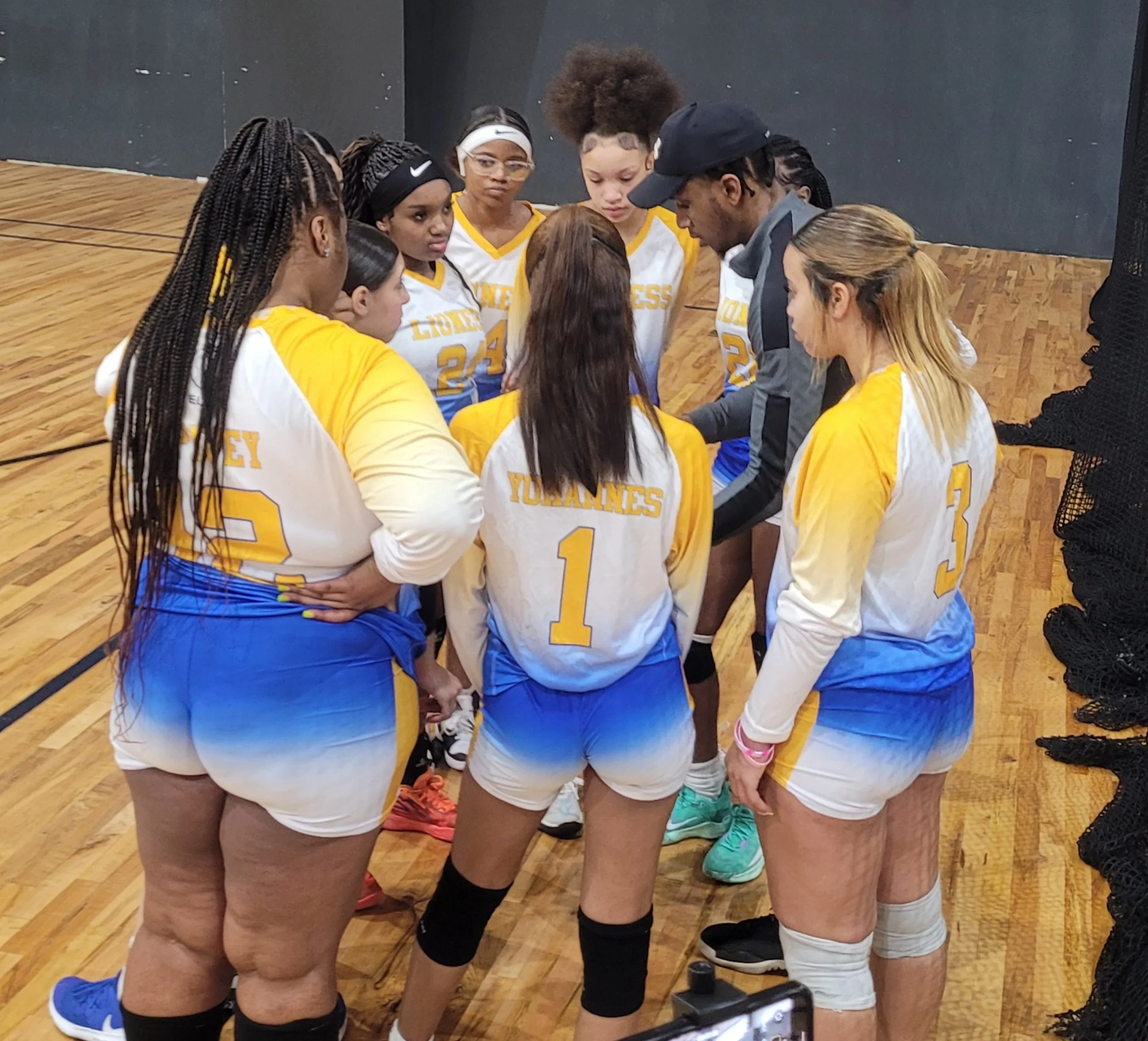 A women's volleyball team in yellow and white jerseys huddles with their coach during a game or practice on a wooden court.