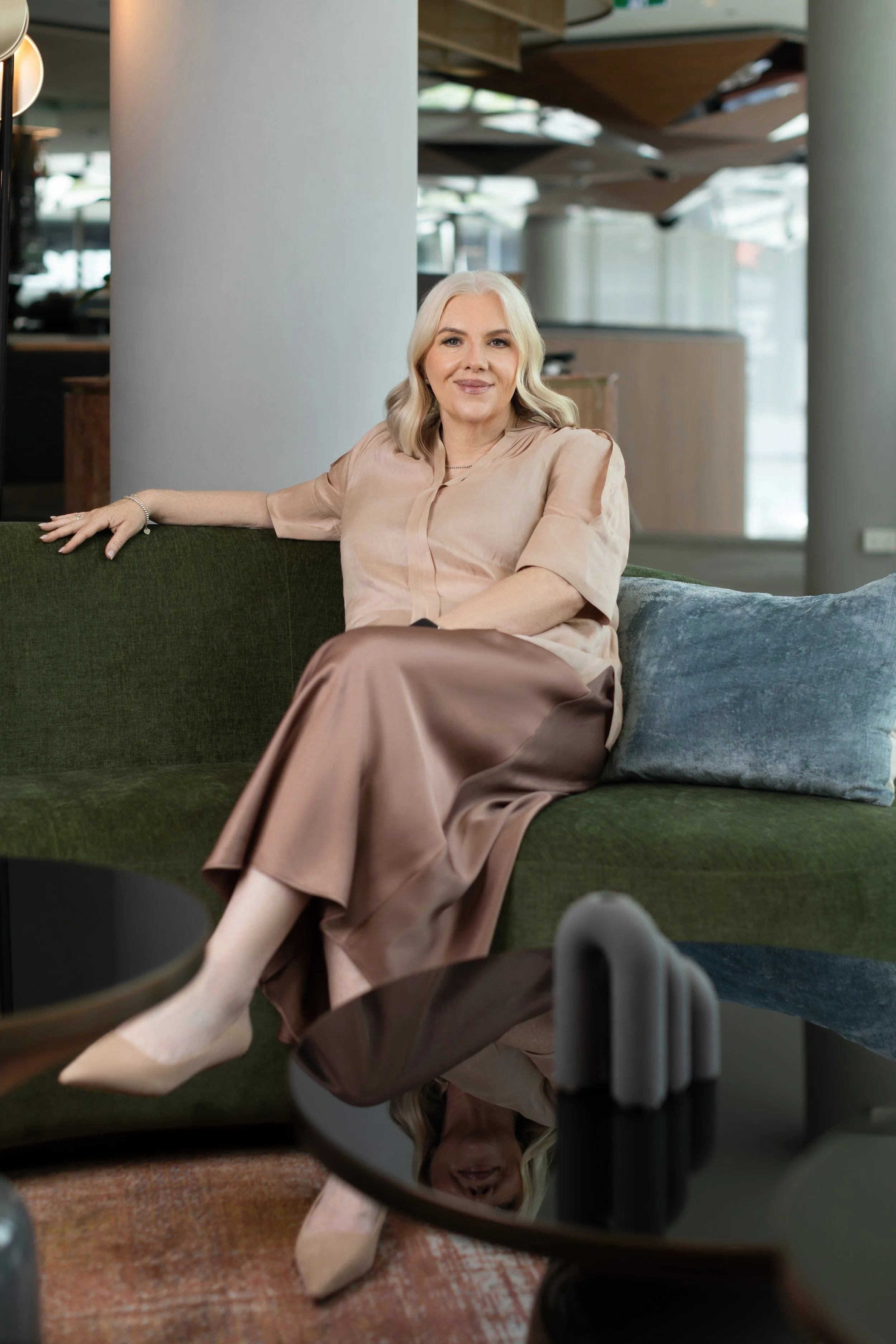 A woman with shoulder-length blonde hair sitting on a green couch in a modern, stylish interior. She is wearing a beige satin blouse, a matching satin skirt, and beige heels. She has a pleasant expression and relaxed posture, with her right arm resti