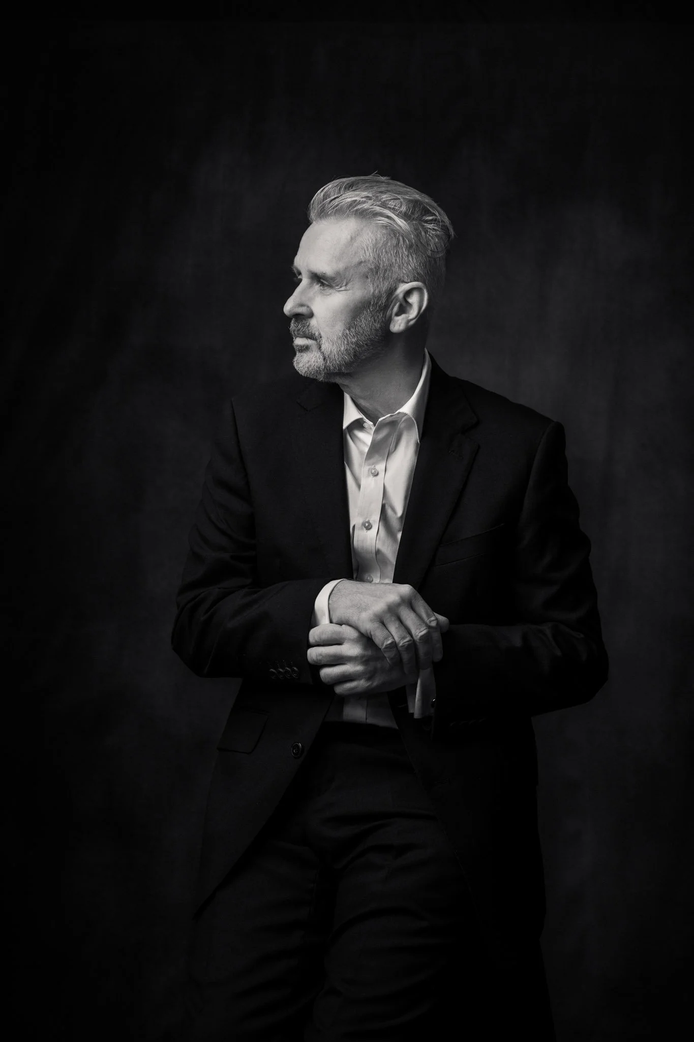 Black and white studio portrait of a well dressed man seated with hands gently wrapped together, looking thoughtfully to the side in a Western Sydney portrait studio