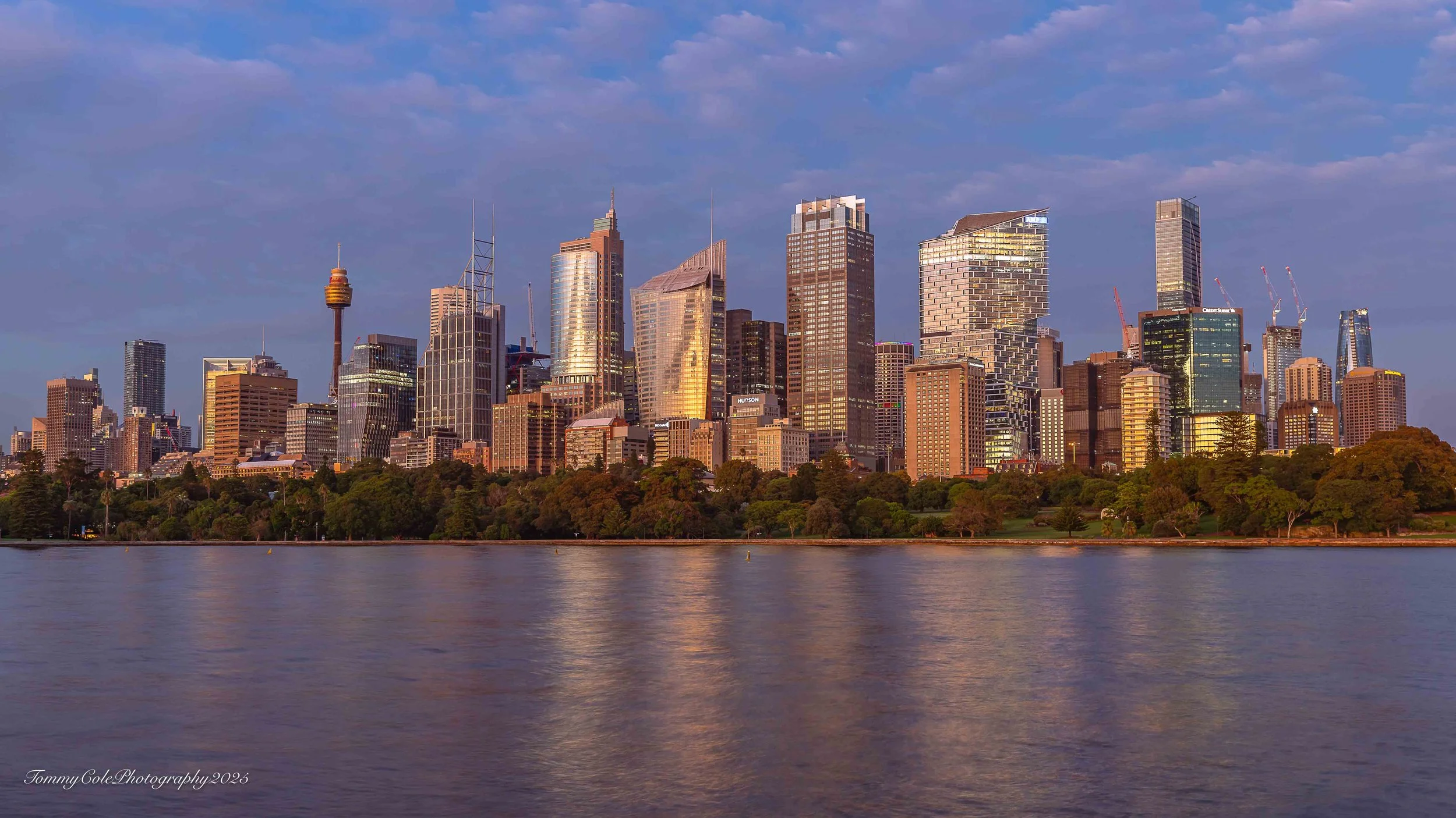 View of downtown skyline reflecting on a river at sunset with a mix of modern skyscrapers and green trees in foreground.
