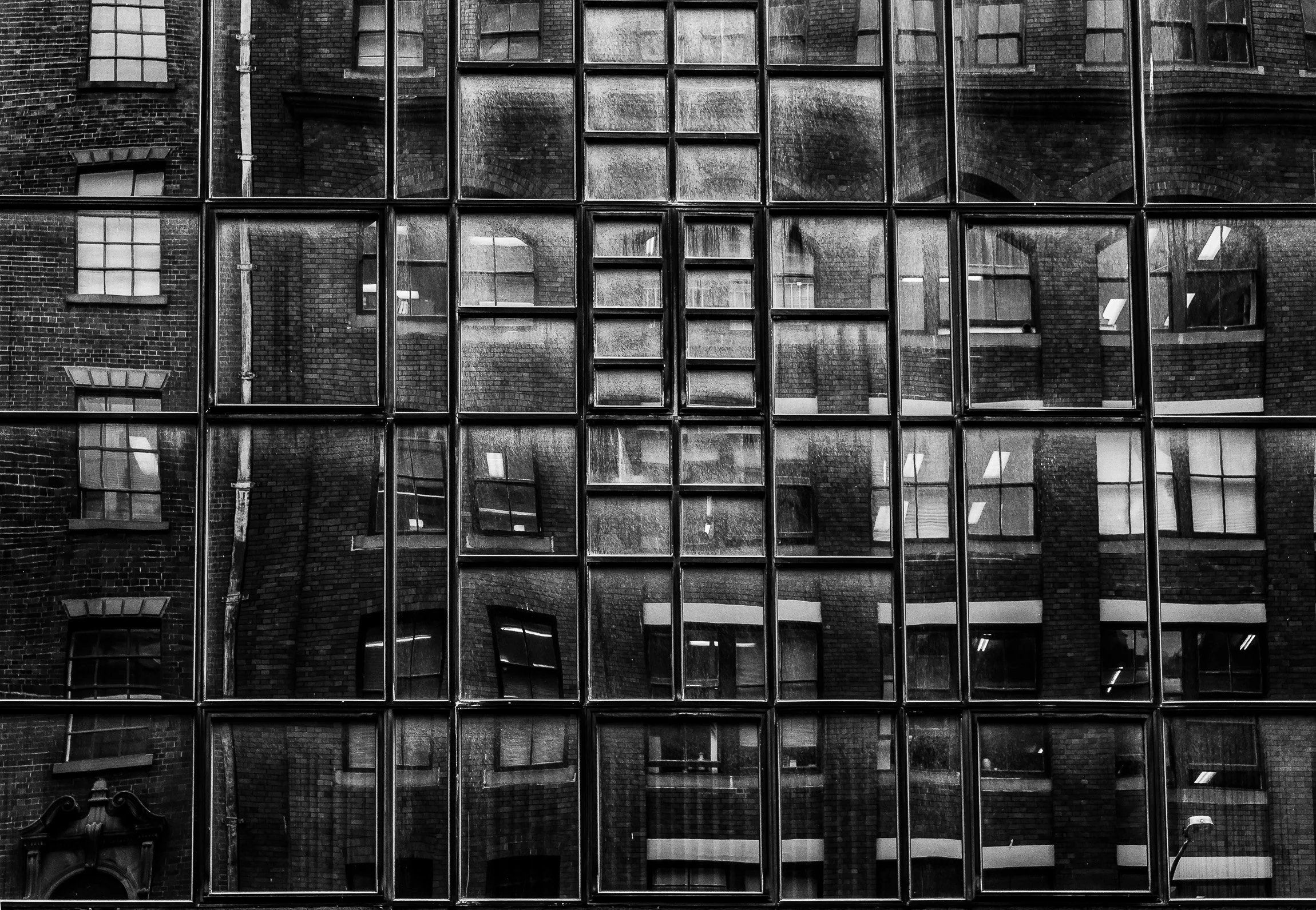 Black and white photo of a building with a reflective glass facade, showing reflections of surrounding brick buildings and windows.