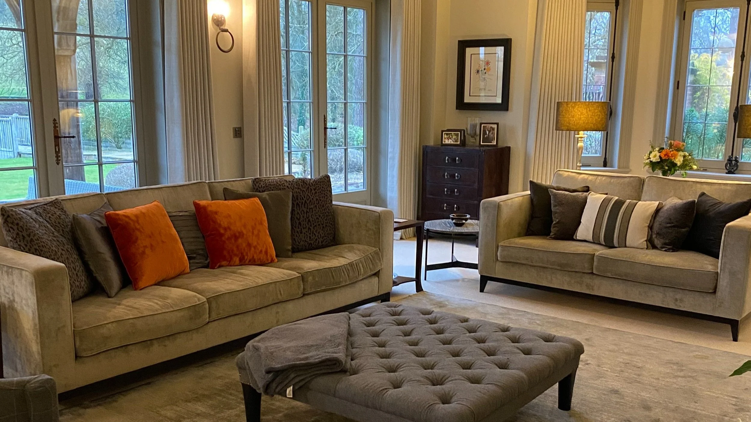 Living room with two beige sofas, a tufted ottoman, and large windows with curtains showing a garden outside. Decor includes colorful pillows, lamps, framed artwork, and a small side table.