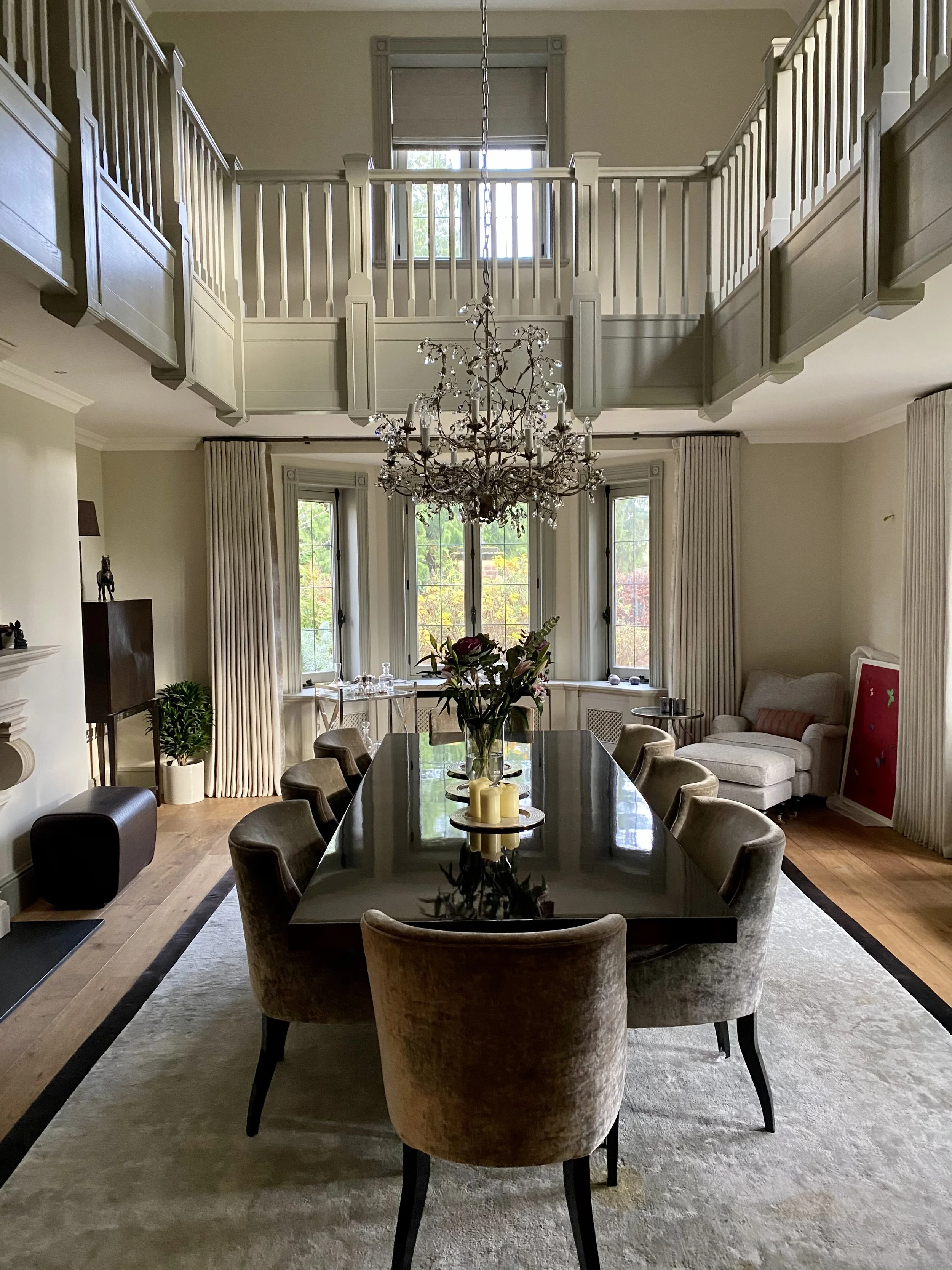 A dining room with a large dark wood table surrounded by beige upholstered chairs, a chandelier hanging above, and large windows with curtains allowing natural light, with a second-floor balcony visible overhead.