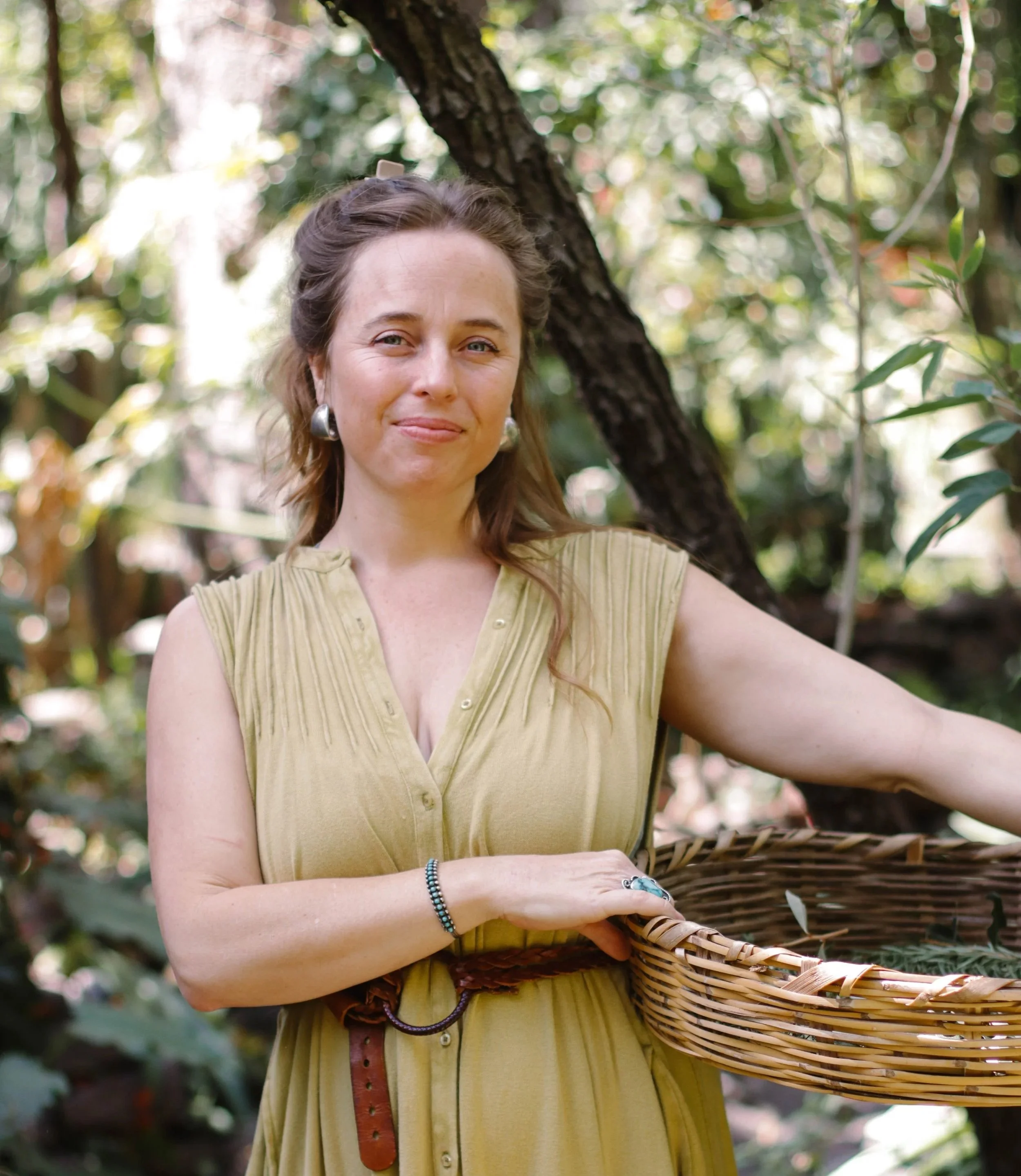 A woman with light skin and brown hair standing outdoors in a forested area, holding a wicker basket, wearing a yellow sleeveless blouse, earrings, and a bracelet.