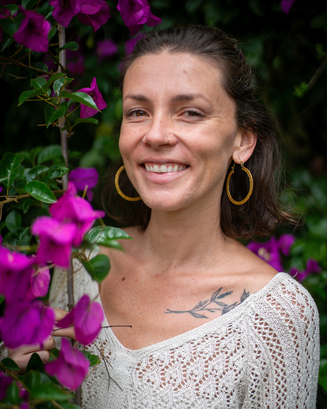 A smiling woman with brown hair, gold hoop earrings, and a tattoo on her shoulder, standing next to purple flowers with green leaves.