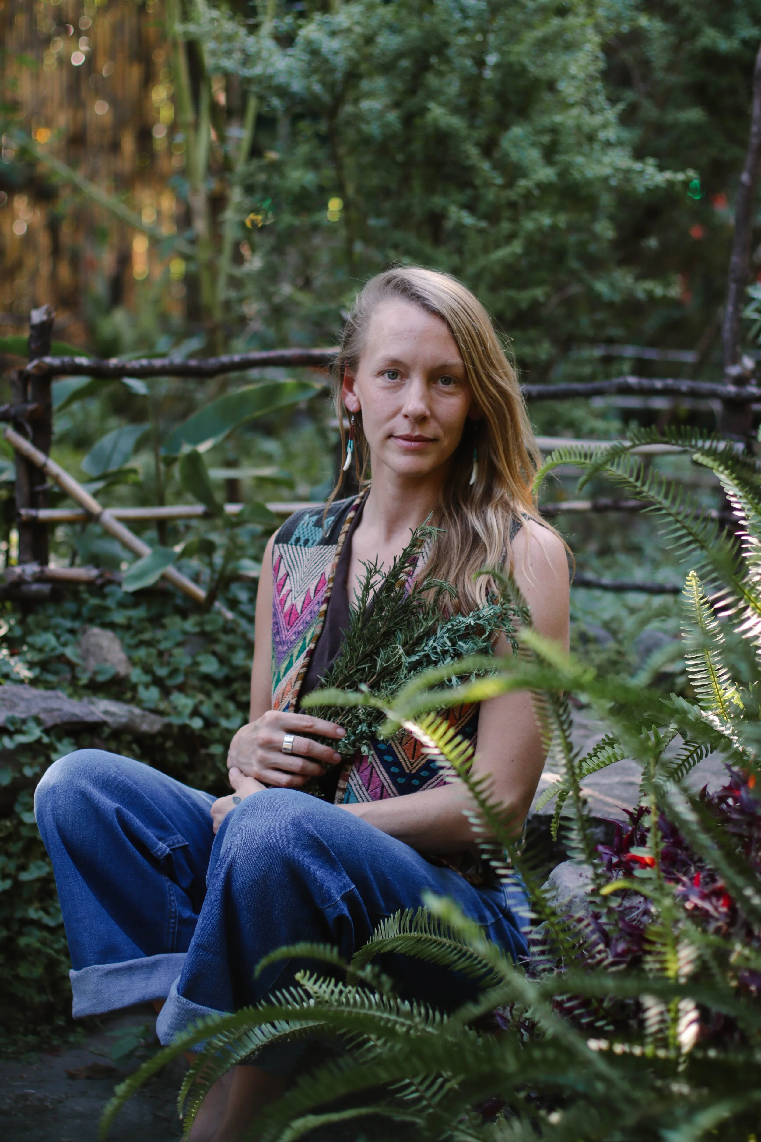 A woman with long, blonde hair and blue eyes sitting outdoors among lush green plants and trees, holding a bunch of greenery in her hands, wearing a colorful patterned sleeveless top and blue jeans.
