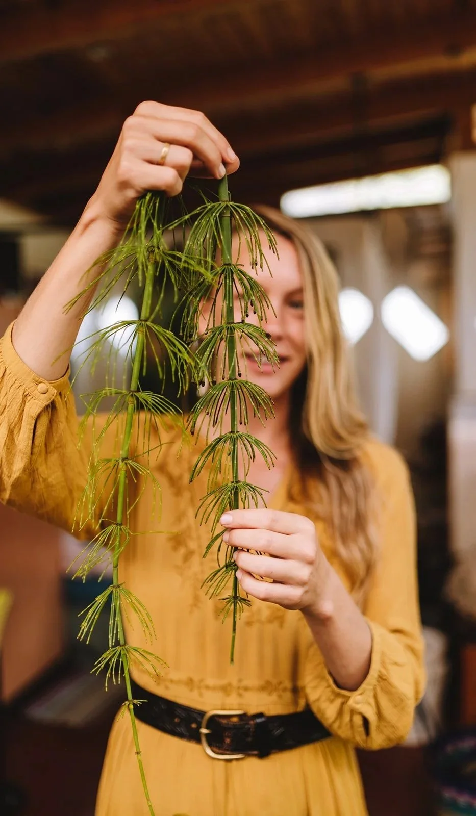 A woman in a yellow dress holding a hanging green plant indoors.